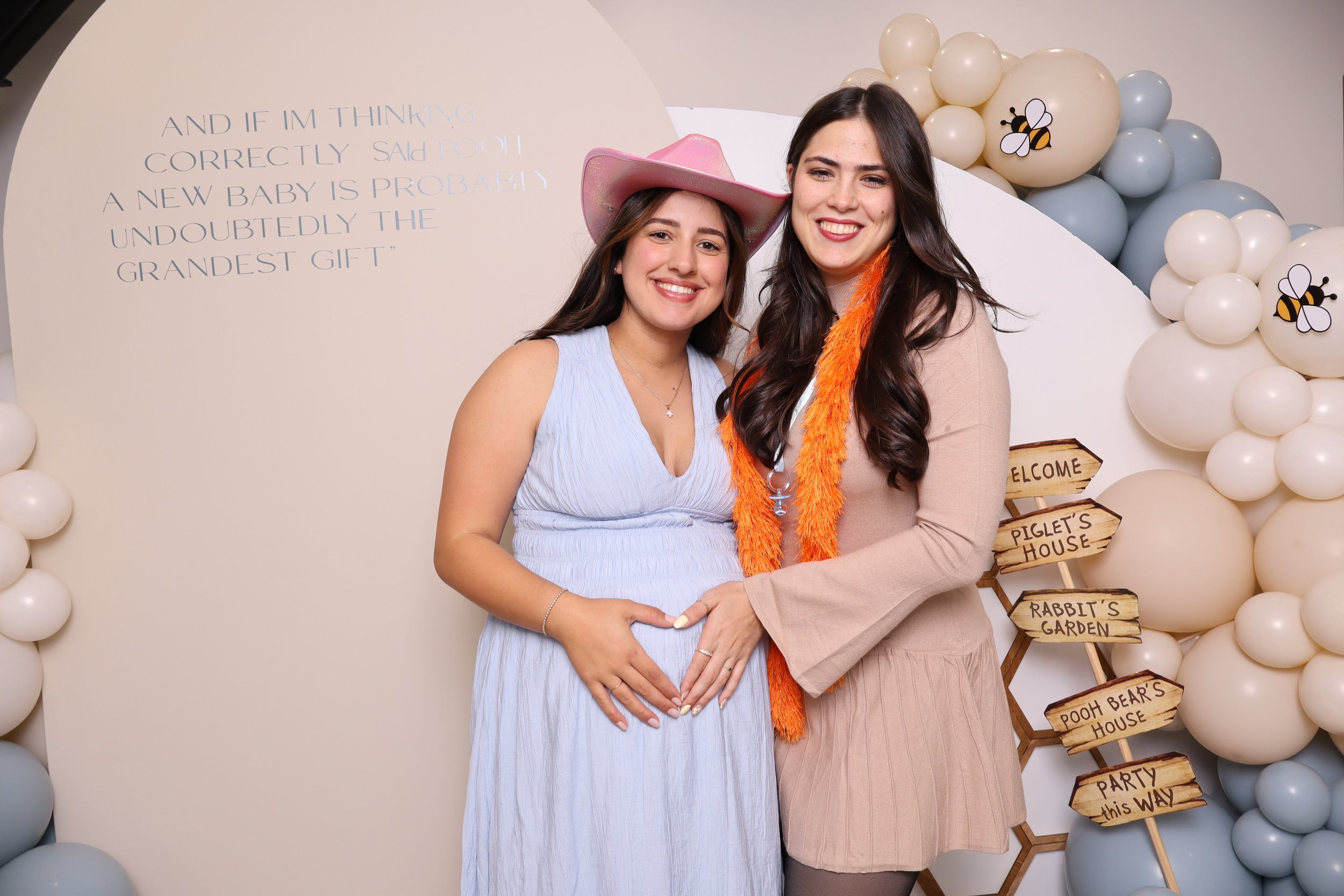 Two smiling women, one pregnant, at a baby shower with balloon decorations and a sign pointing to various themed rooms like Piglet's House and Winnie the Pooh's House.