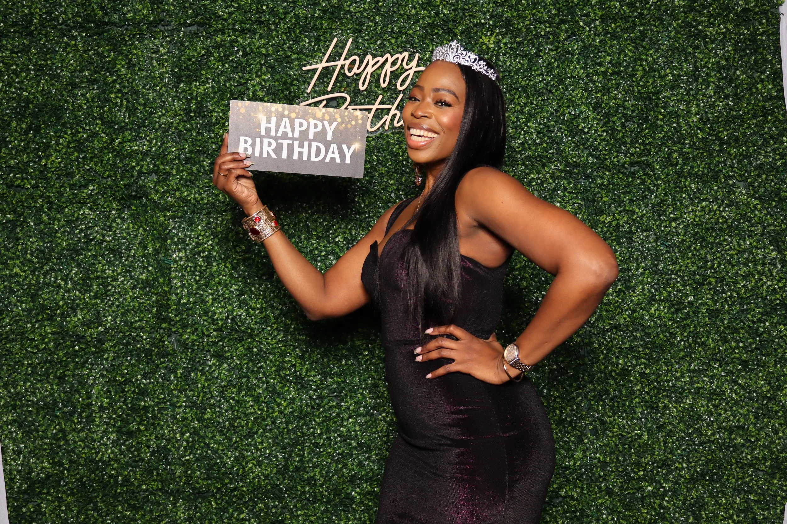 Woman wearing a tiara holding a 'Happy Birthday' sign, standing in front of a green hedge backdrop with a 'Happy Birthday' sign on the wall.