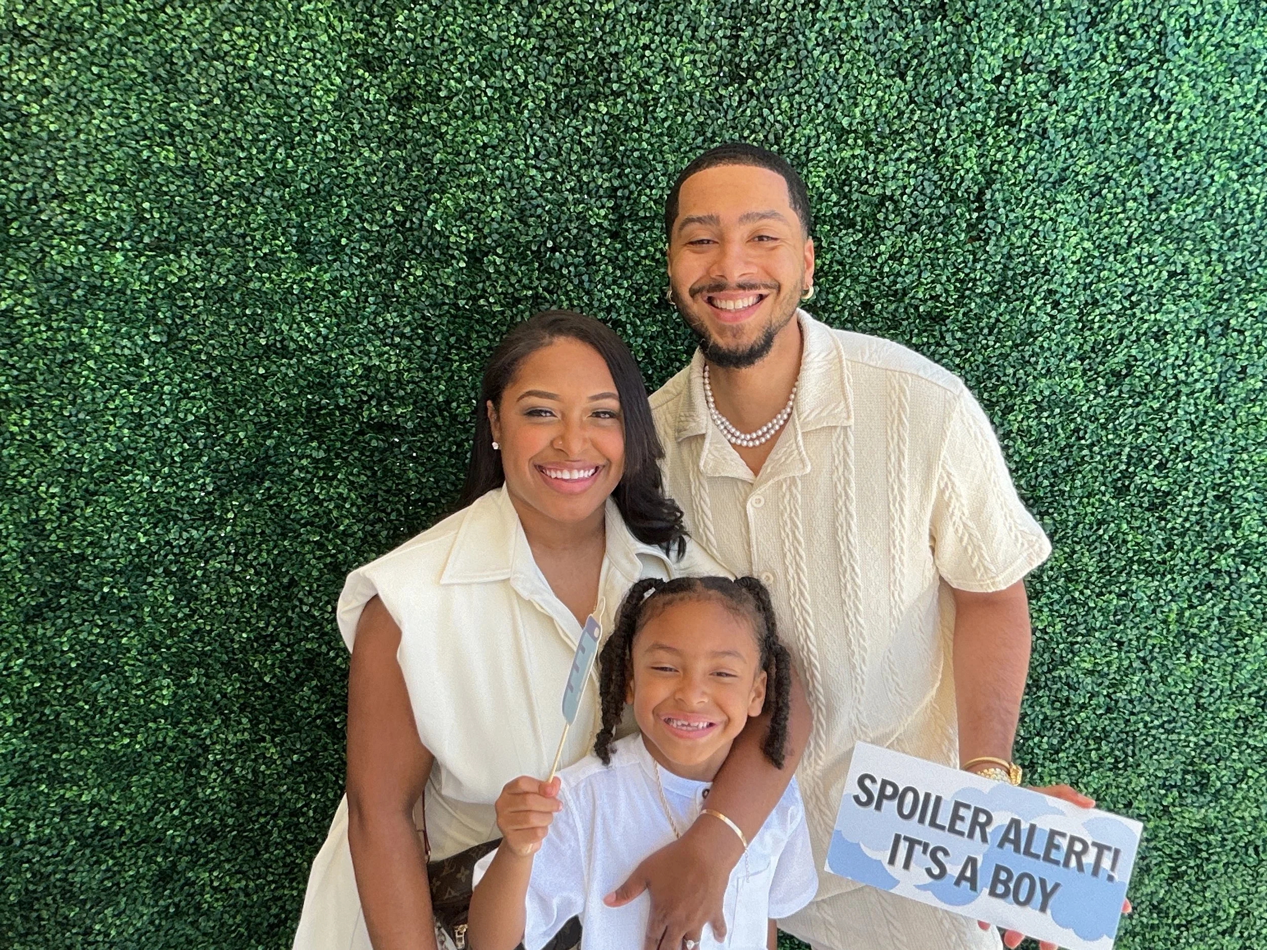 Family of three celebrating, holding a sign that reads 'Spoiler Alert! It's a Boy'. They are standing in front of a green leafy background, all smiling, dressed in light-colored clothing.
