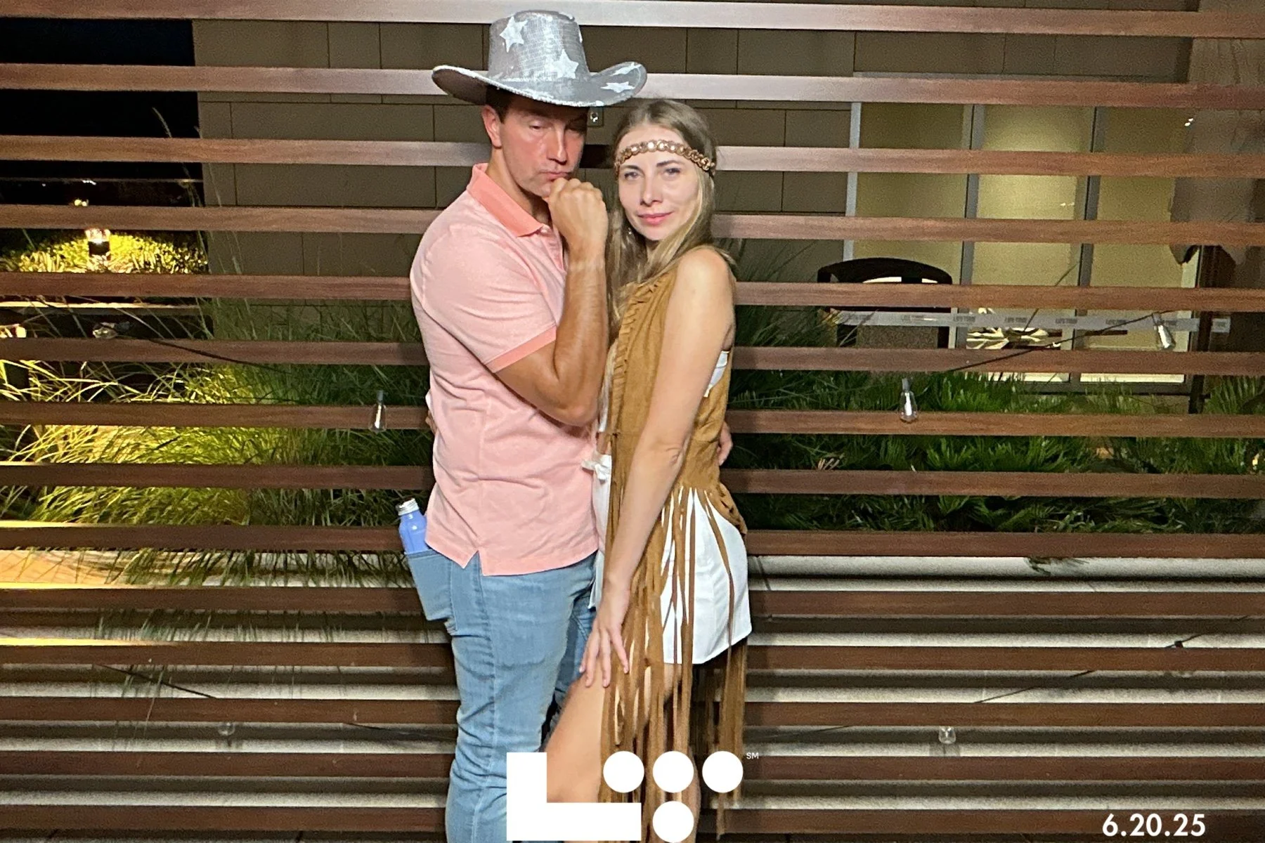 A man wearing a pink shirt, jeans, and a star-patterned cowboy hat stands next to a woman in an outfit with fringes and a headband, posing in front of a wooden slat wall at night.