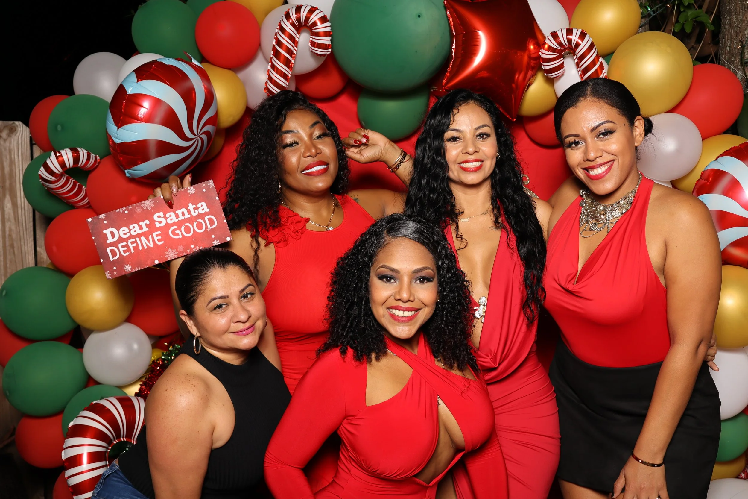 Five women celebrating Christmas in front of a colorful balloon backdrop, with one woman holding a sign that reads 'Dear Santa, DEFINE GOOD'.