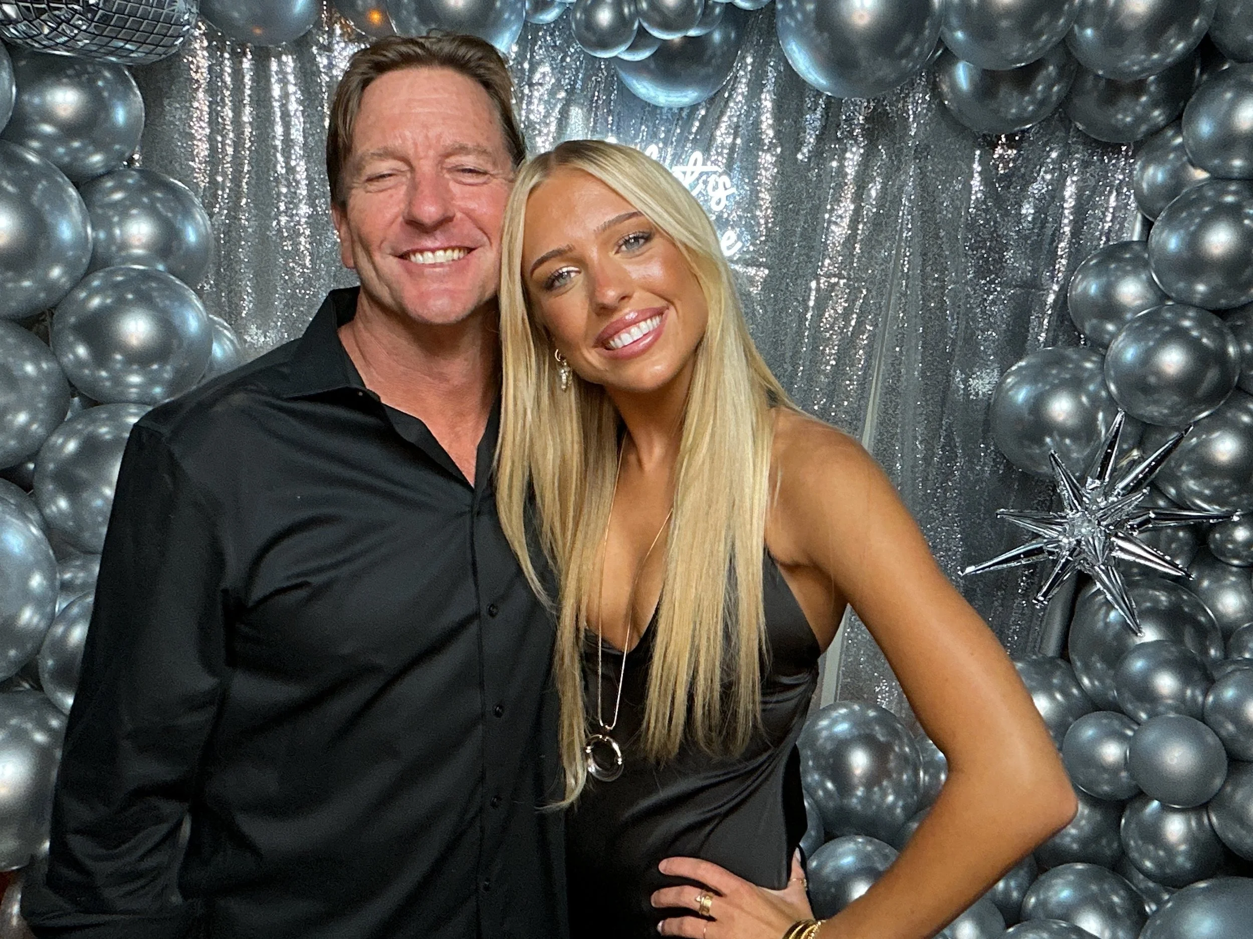 A smiling man and woman posing together in front of a silver balloon backdrop with a glittery curtain. The man is wearing a black button-up shirt, and the woman is wearing a black sleeveless dress.