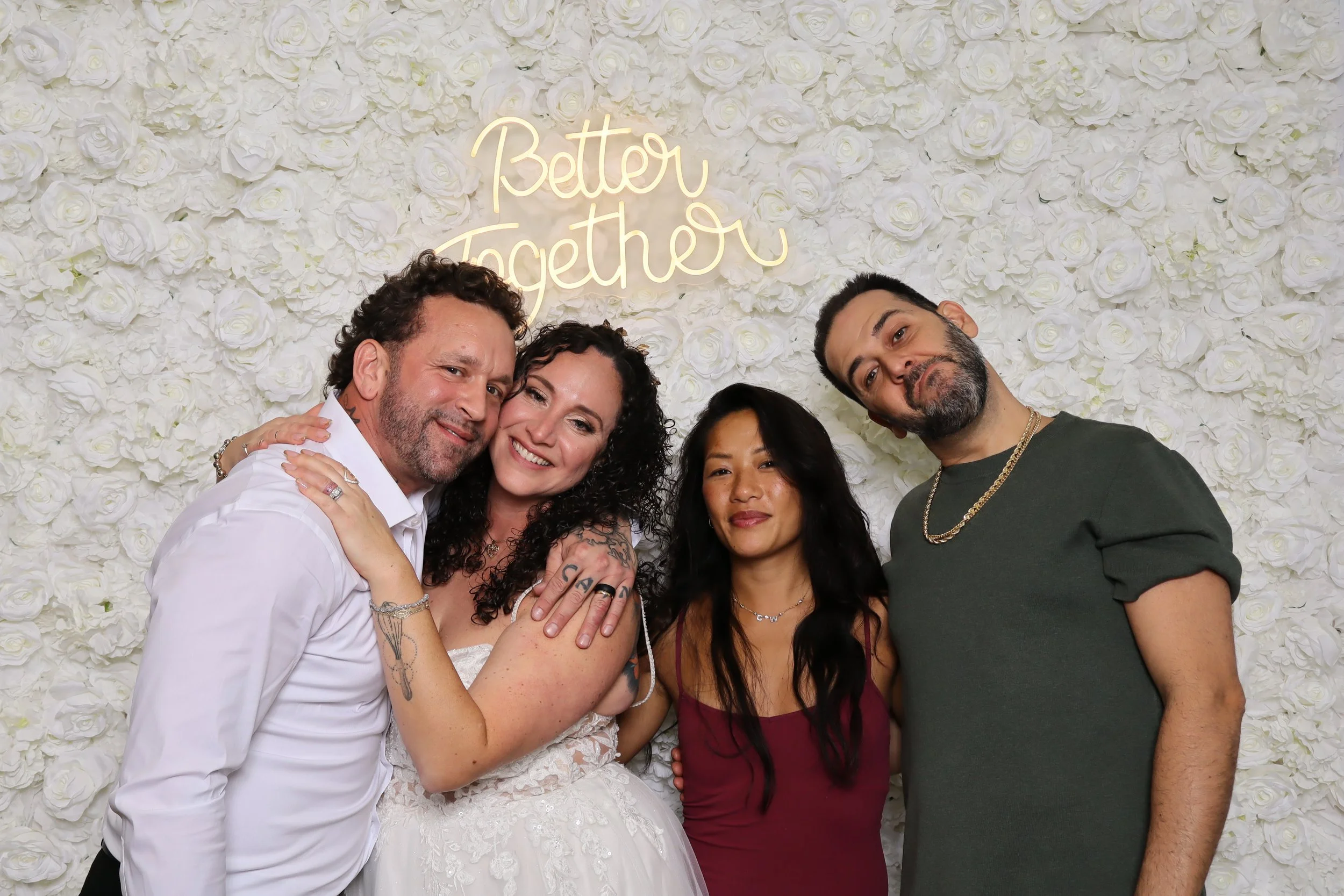 Four people smiling and posing for a photo in front of a white rose wall with a neon sign that says 'Better Together.'
