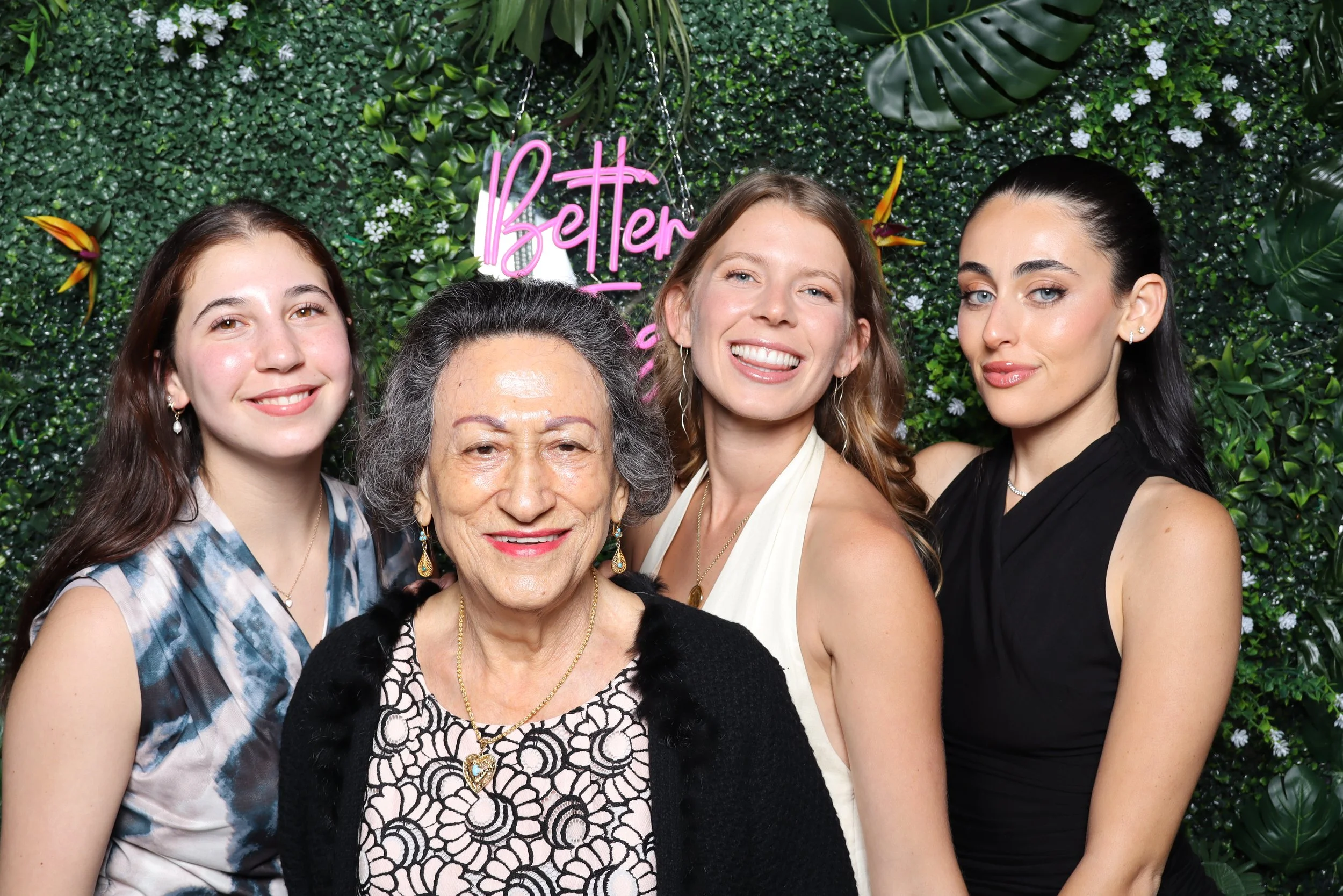 Four women of different ages smiling and posing together in front of a green leafy background with a neon pink sign that says 'Better'.