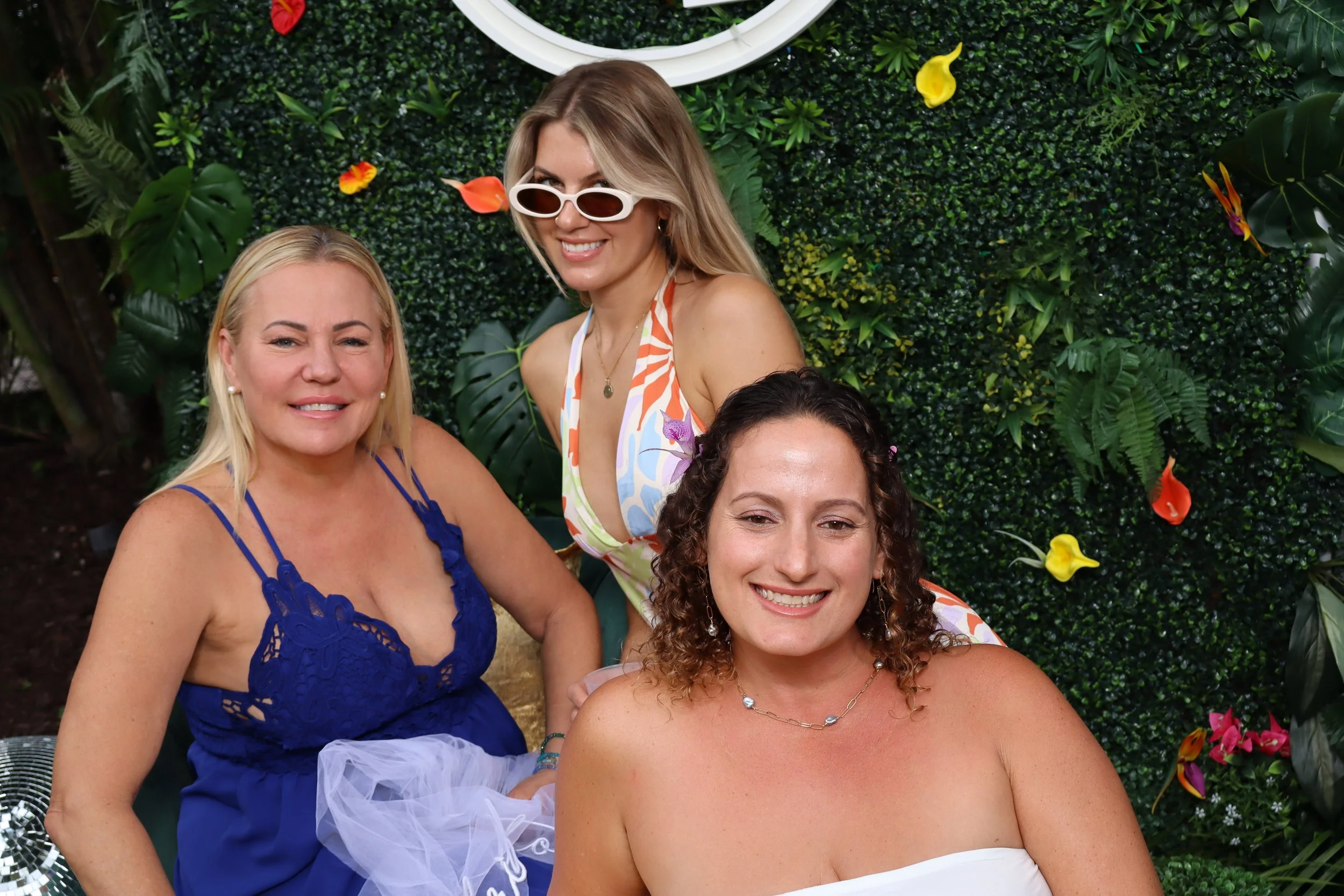 Three women smiling at a garden party with a green leafy backdrop and colorful flowers, one wearing a blue dress, another in a multicolored halter top, and the third in a white strapless dress.