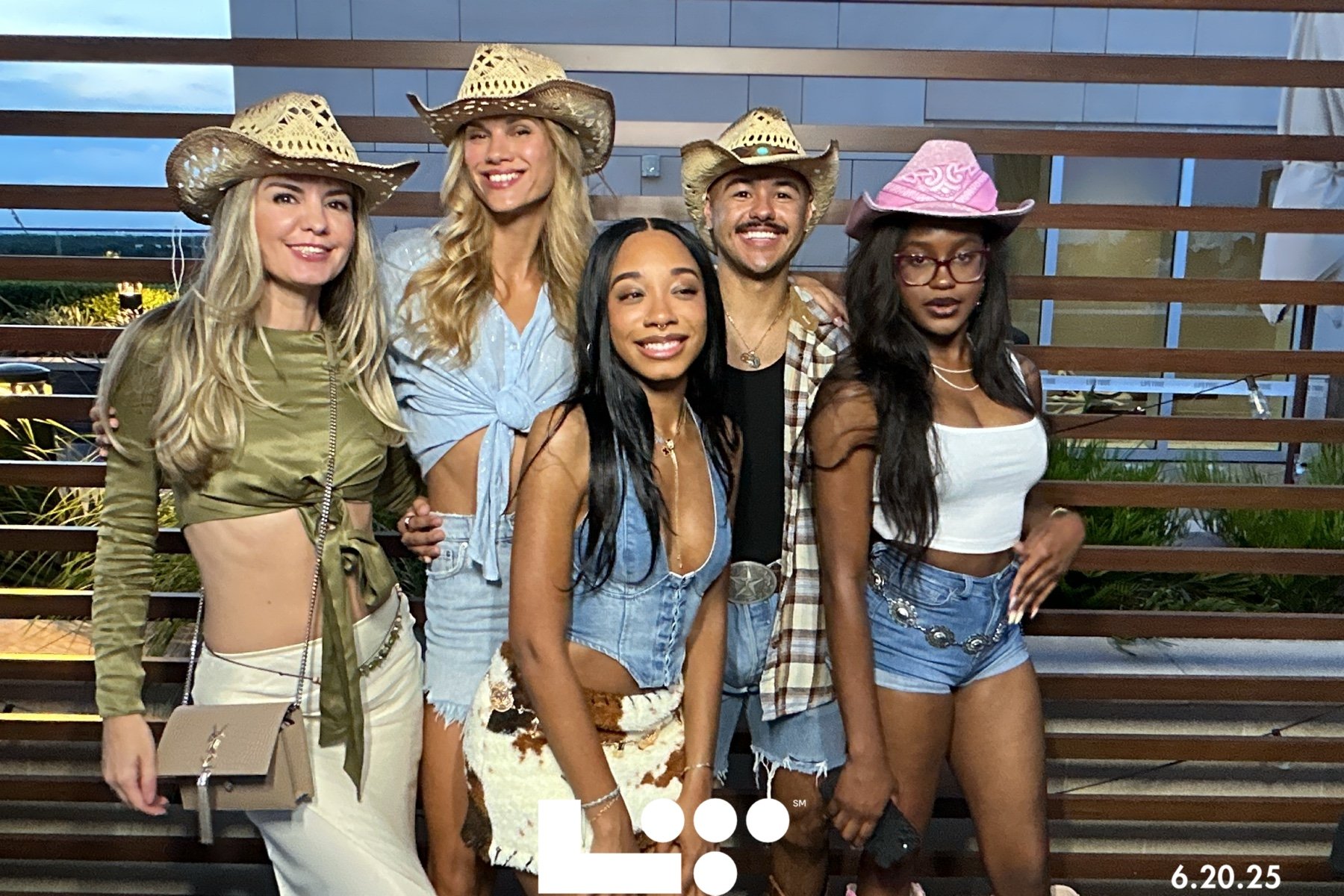 Five young adults, three women and two men, dressed in casual summer outfits with cowboy hats, posing outdoors on a balcony with wooden slats and greenery behind them, during evening.