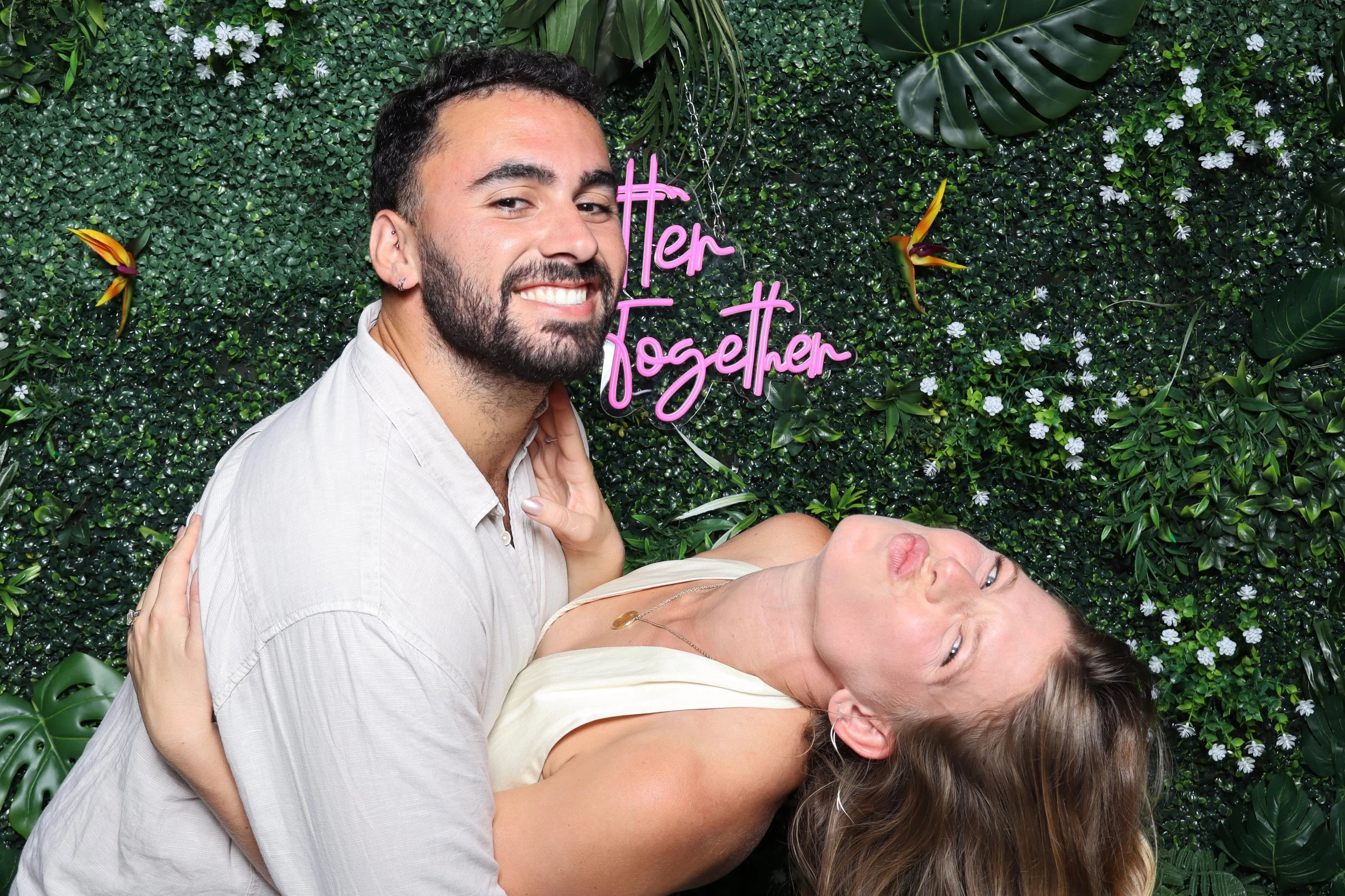 Two smiling people, a man and a woman, pose in front of a green leafy wall with a neon pink sign that reads 'Terk Together'. The woman is leaning back with her head tilted and the man is smiling at the camera.