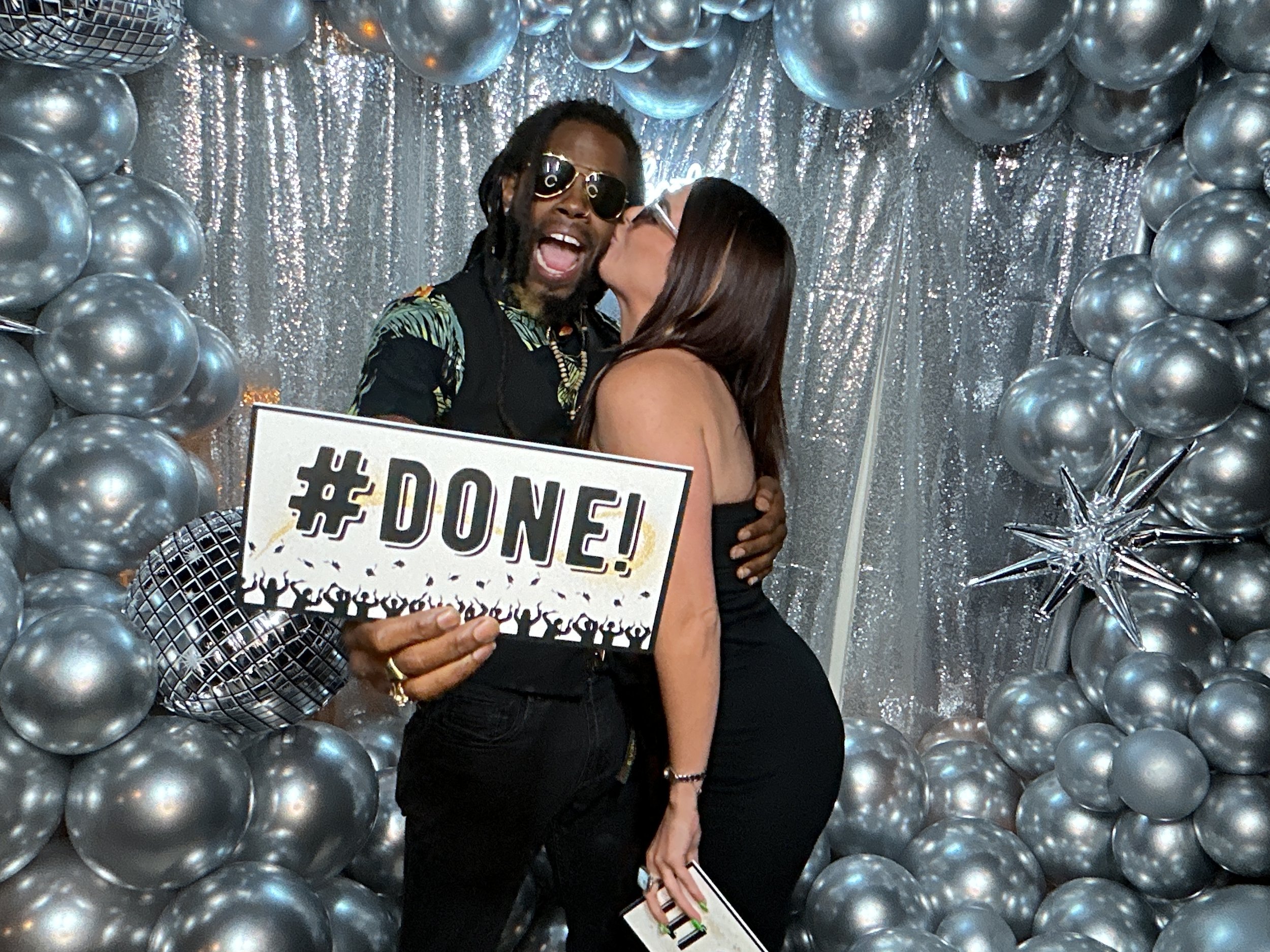Two people celebrating at a party with silver balloons and a shiny backdrop; one person is holding a sign that says '#DONE!' and hugging the other person, who is giving a kiss on the cheek.