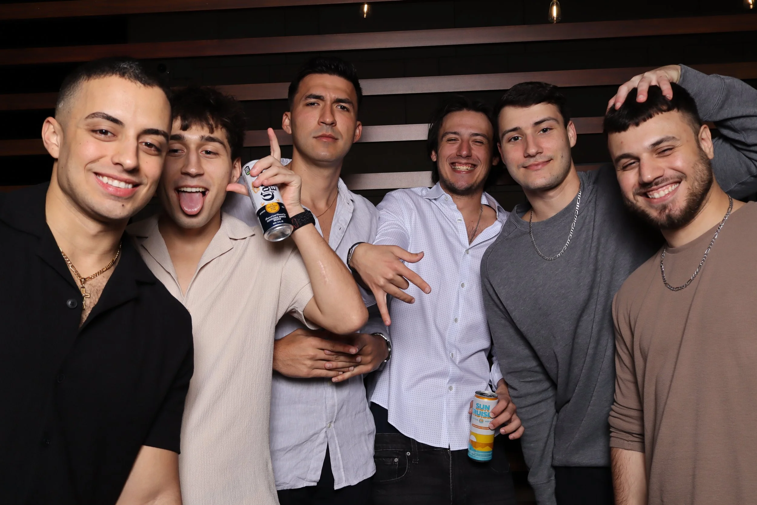 Group of seven young men posing together at a party, smiling and making gestures, with some holding drinks, against a dark wooden background.