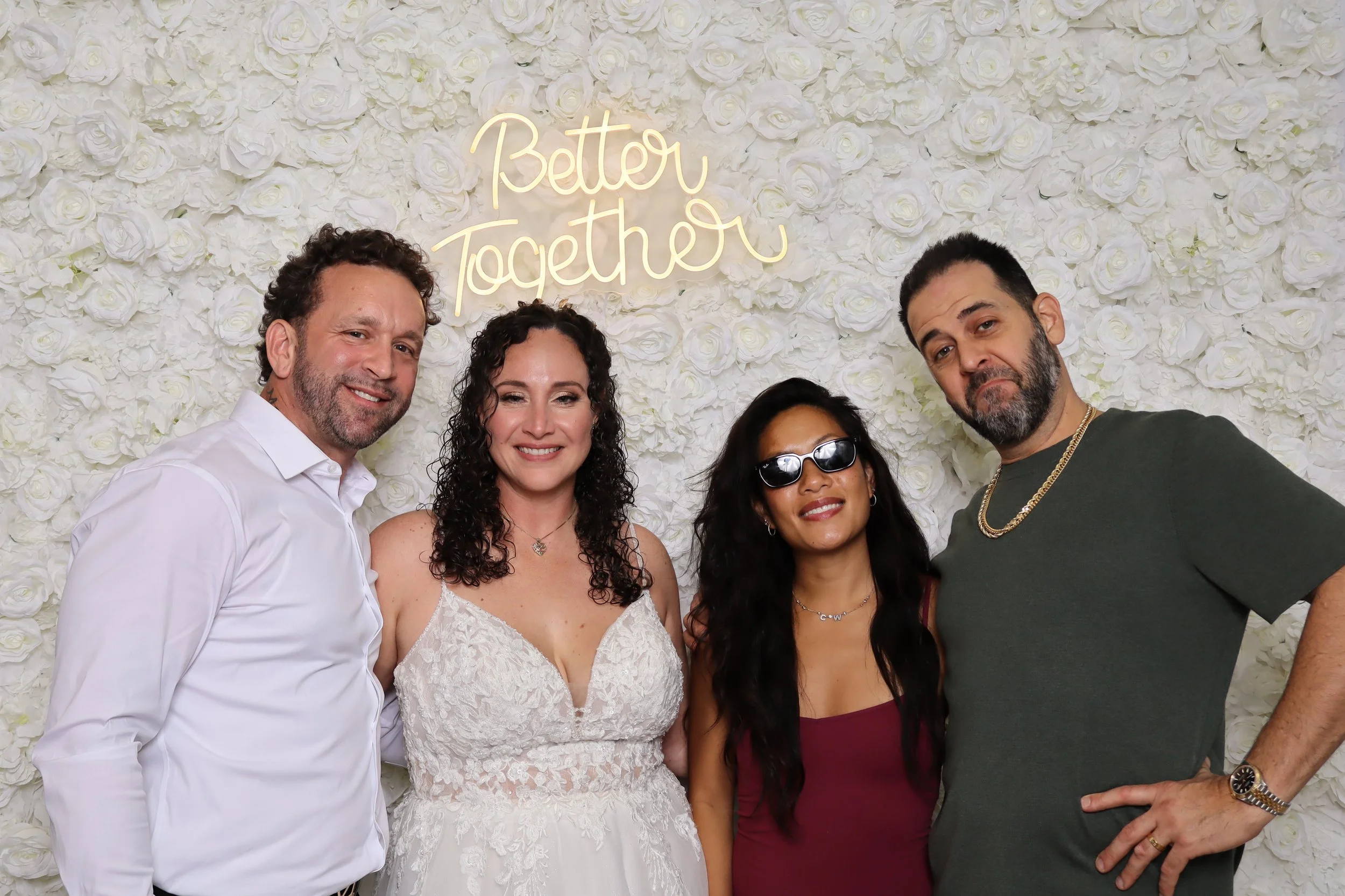 Four people standing in front of a white flower wall, with a neon sign that says 'Better Together' in cursive. The group includes two men and two women, all smiling, with one woman wearing sunglasses.