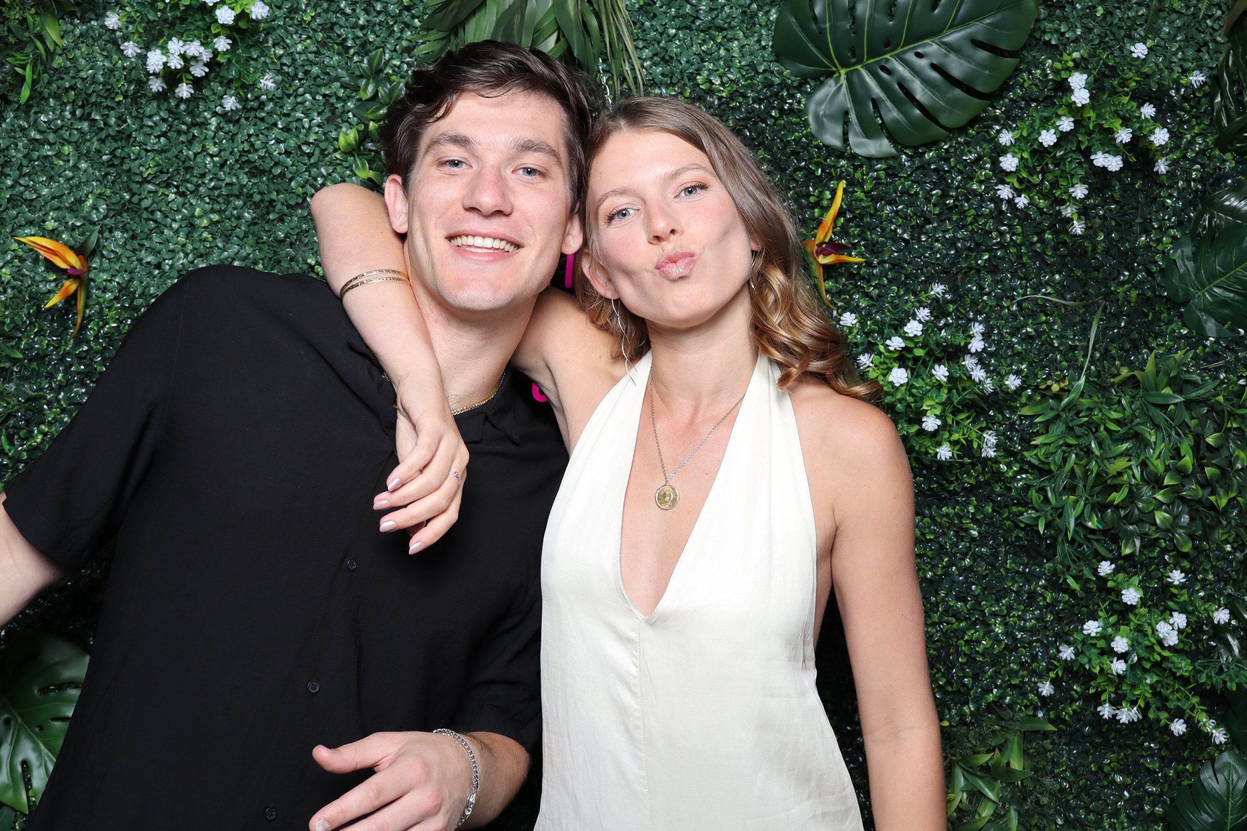 A young man and woman are posing closely together, smiling and making a kissing face in front of a green leafy floral background.