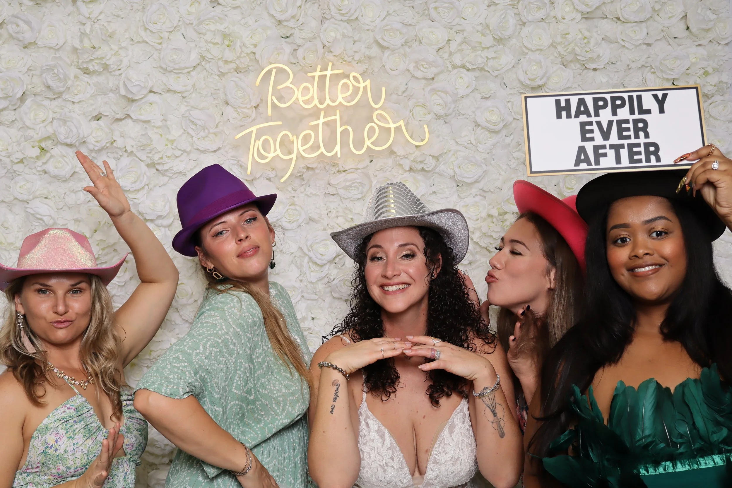 Group of five women wearing colorful hats, smiling and posing in front of a white floral backdrop with neon sign that says 'Better Together,' and a sign that says 'Happily Ever After.'