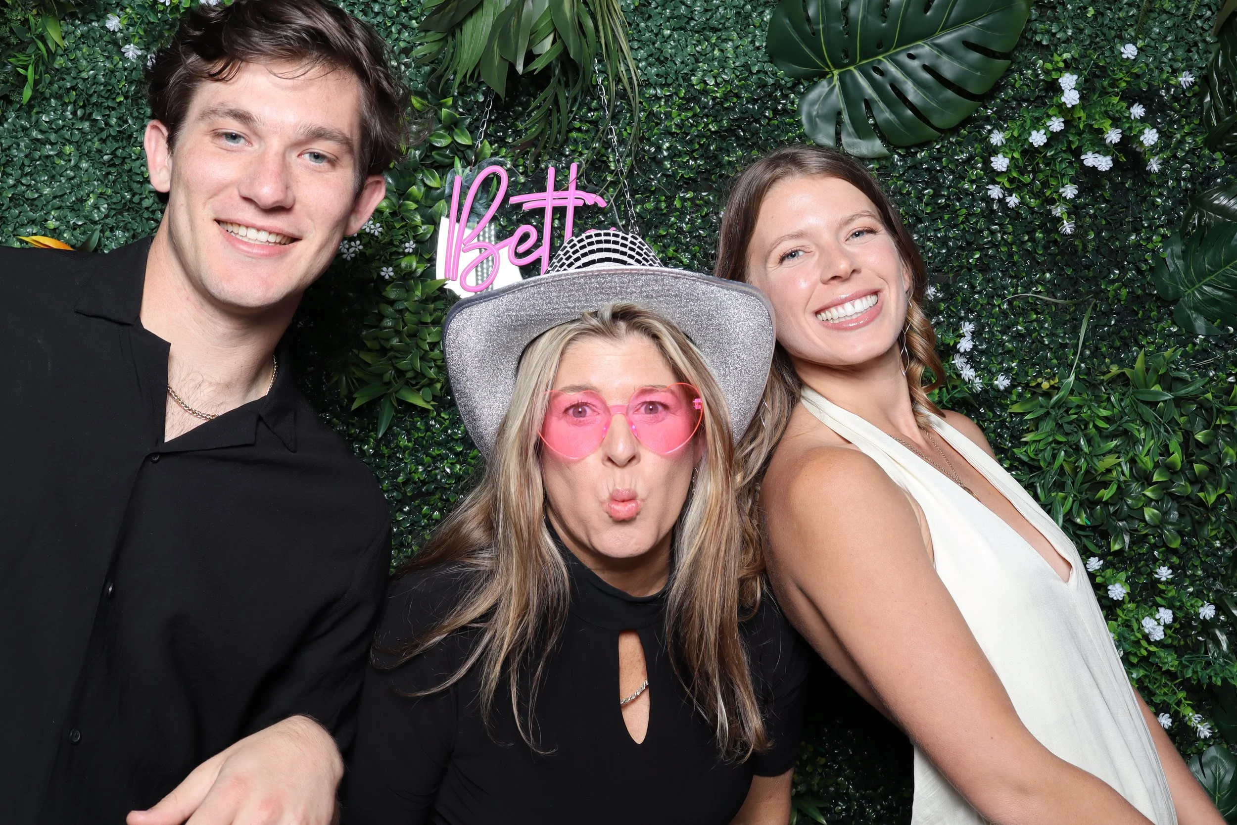 Three people smiling and posing for a photo in front of a green leafy backdrop with a neon pink 'Bet' sign. The person in the middle is wearing pink heart-shaped sunglasses and a large silver hat, while the other two are dressed casually, with one in