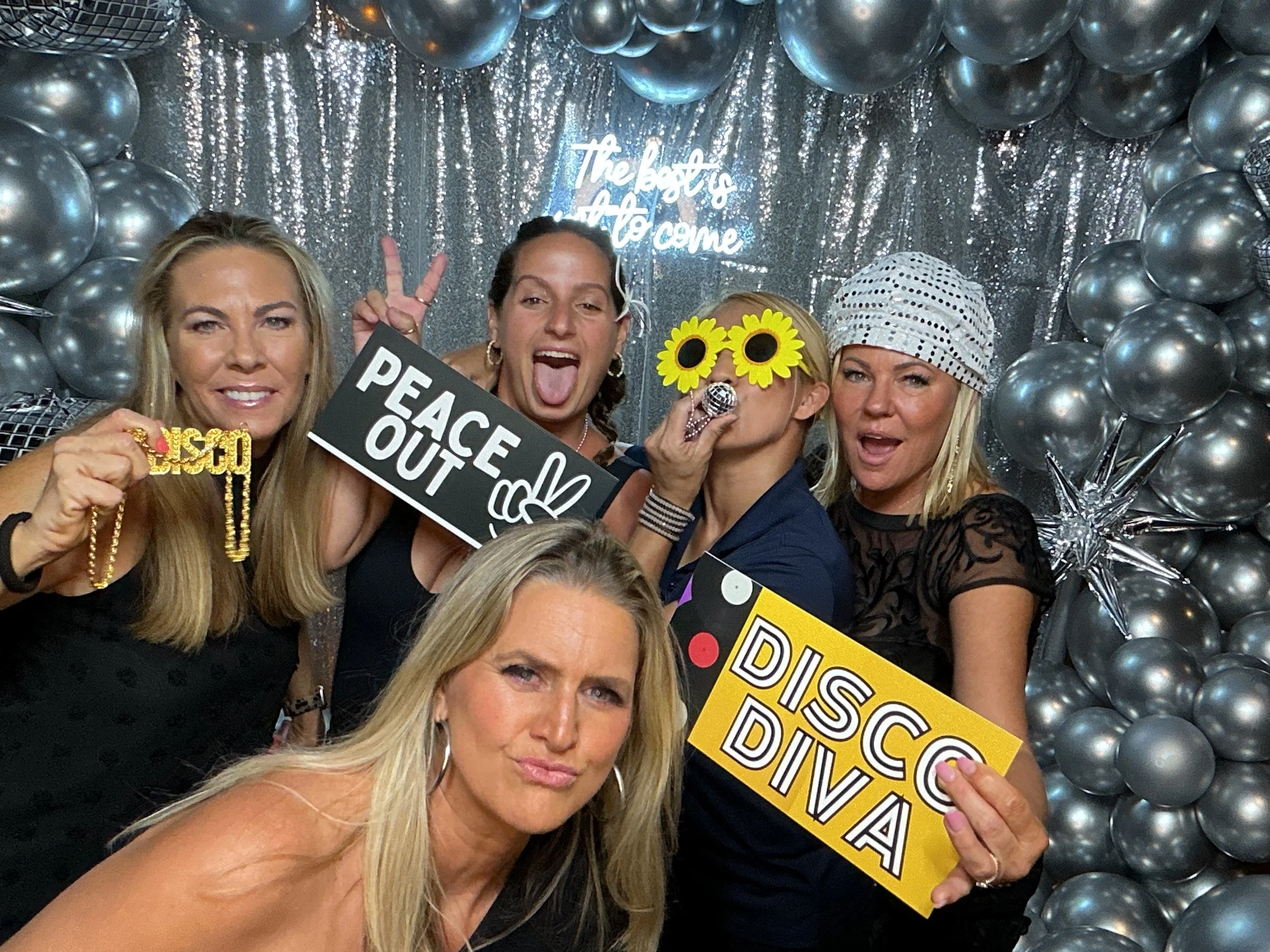 Group of five women at a disco-themed party, surrounded by silver balloons and a sequin backdrop, with signs reading 'Peace Out' and 'Disco Diva', some making playful gestures and wearing fun accessories like sunflower glasses and a glittery hat.