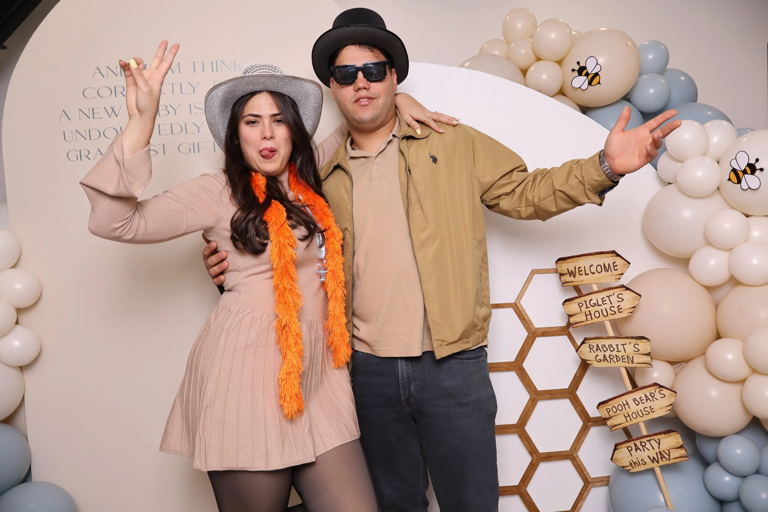 A woman and a man posing at a themed event with honeycomb and bee decorations. The woman is making a peace sign and sticking out her tongue, wearing a beige dress, a gray hat, and an orange feather boa. The man is wearing sunglasses, a black hat, a t