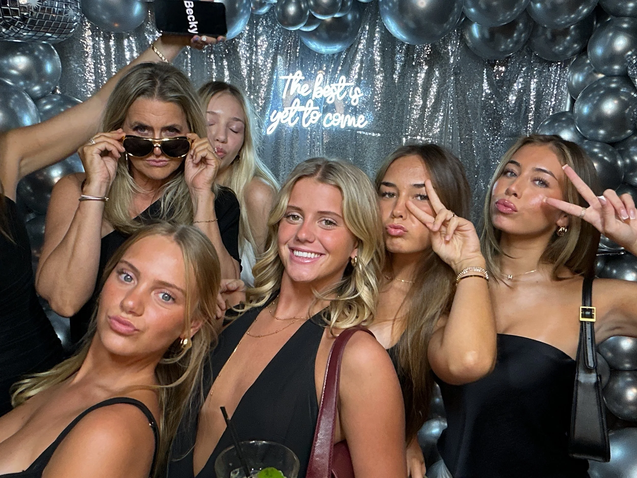 Group of six young women posing for a photo at a party with metallic silver balloons and a silver backdrop, some making peace signs and one woman adjusting sunglasses, with a neon sign that reads 'The best is yet to come' in the background.