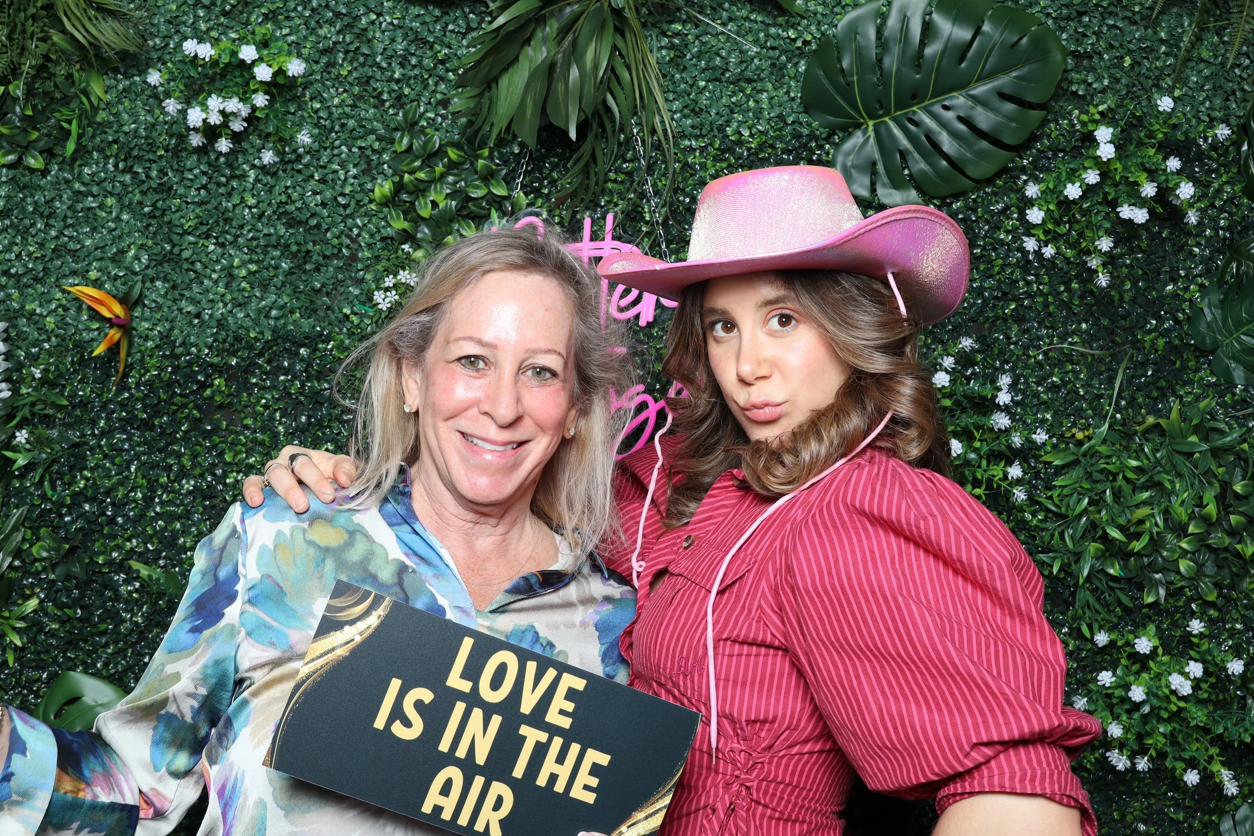 A woman and a young girl posing together in front of a green leafy backdrop. The woman is holding a sign that reads 'Love is in the air.' The girl is wearing a pink cowboy hat and a pink shirt, making a playful pouty face.