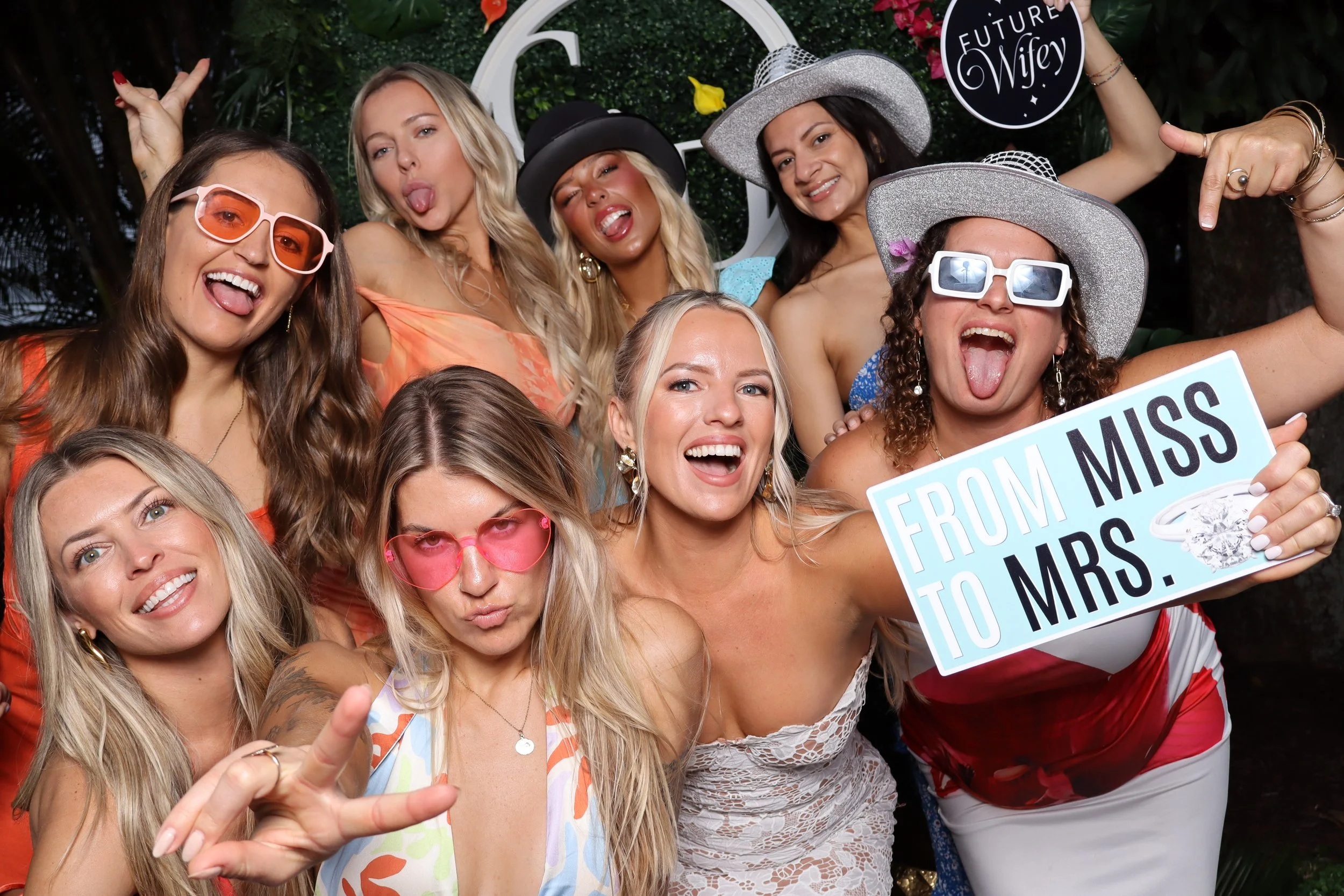 Group of women celebrating a wedding, some wearing colorful dresses and sunglasses, holding a sign that says 'FROM MISS TO MRS.'