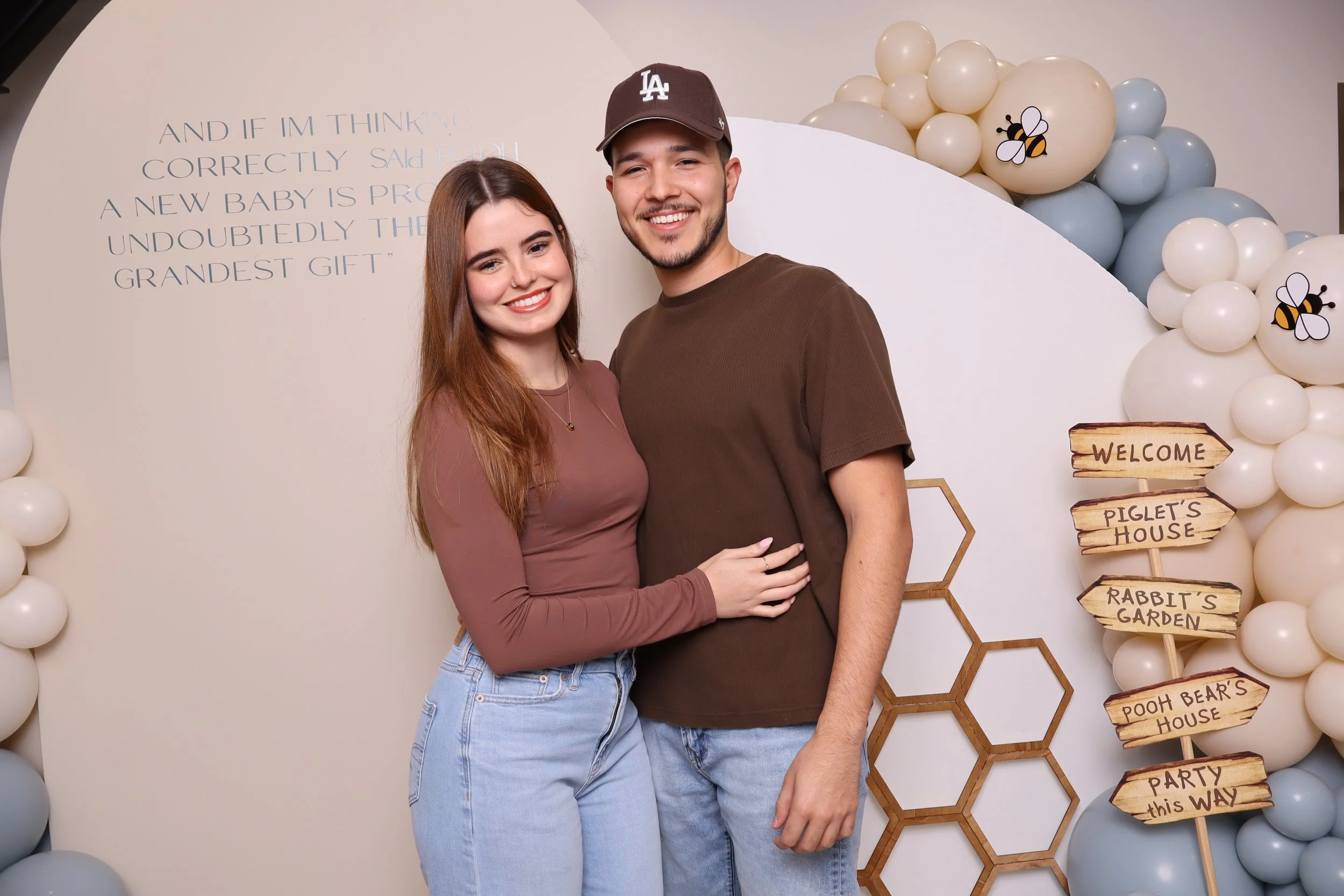 A young couple raises their arms and smiles in front of a white backdrop decorated with balloons and a sign that reads "And if I'm thinking correctly…" with a quote. The woman has long brown hair, wears a mauve long-sleeve top and light blue jeans. T