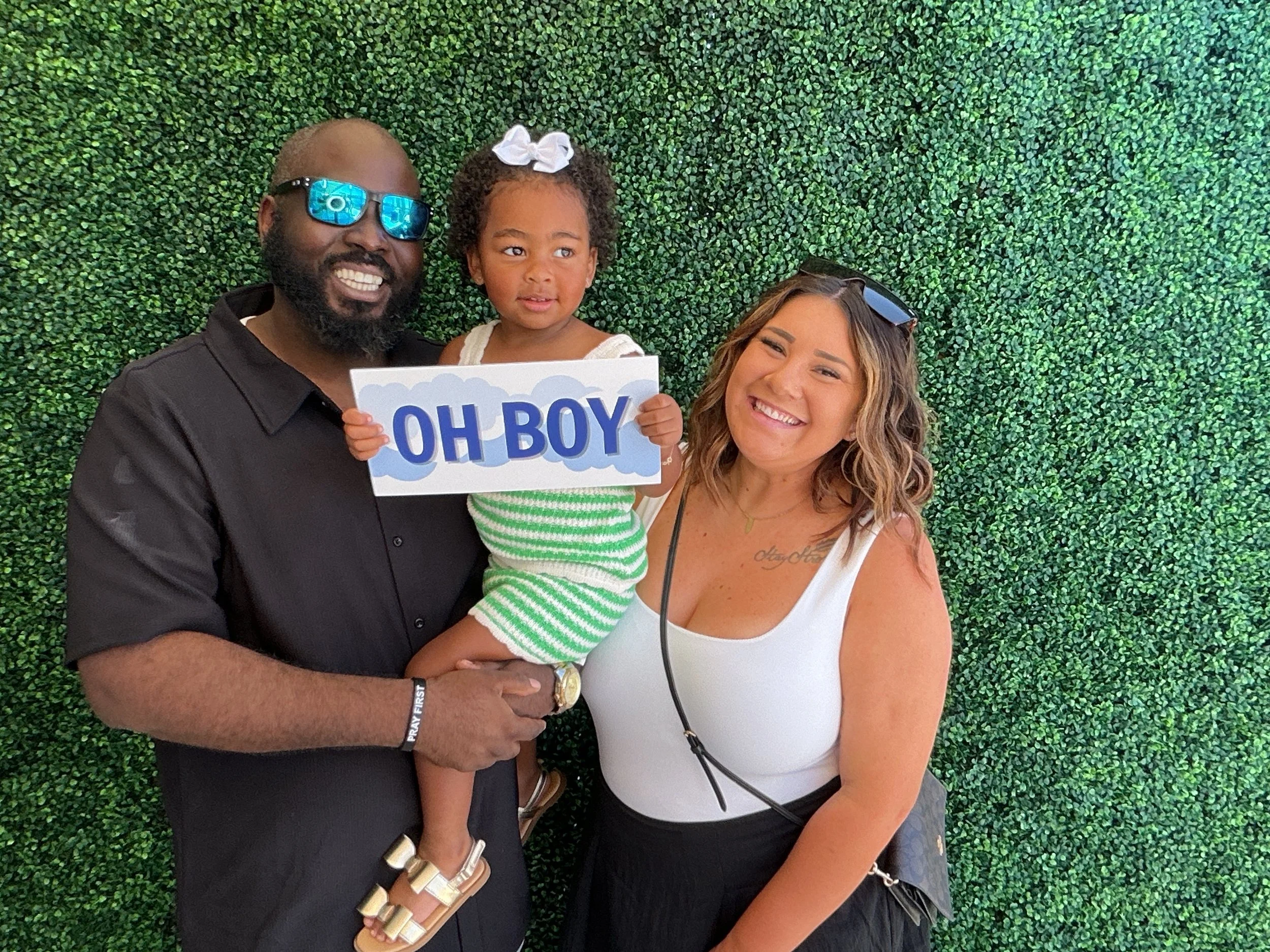 A family of three, including a man with sunglasses, a woman, and a young girl holding a sign that says "OH BOY", standing against a green leafy wall, all smiling.