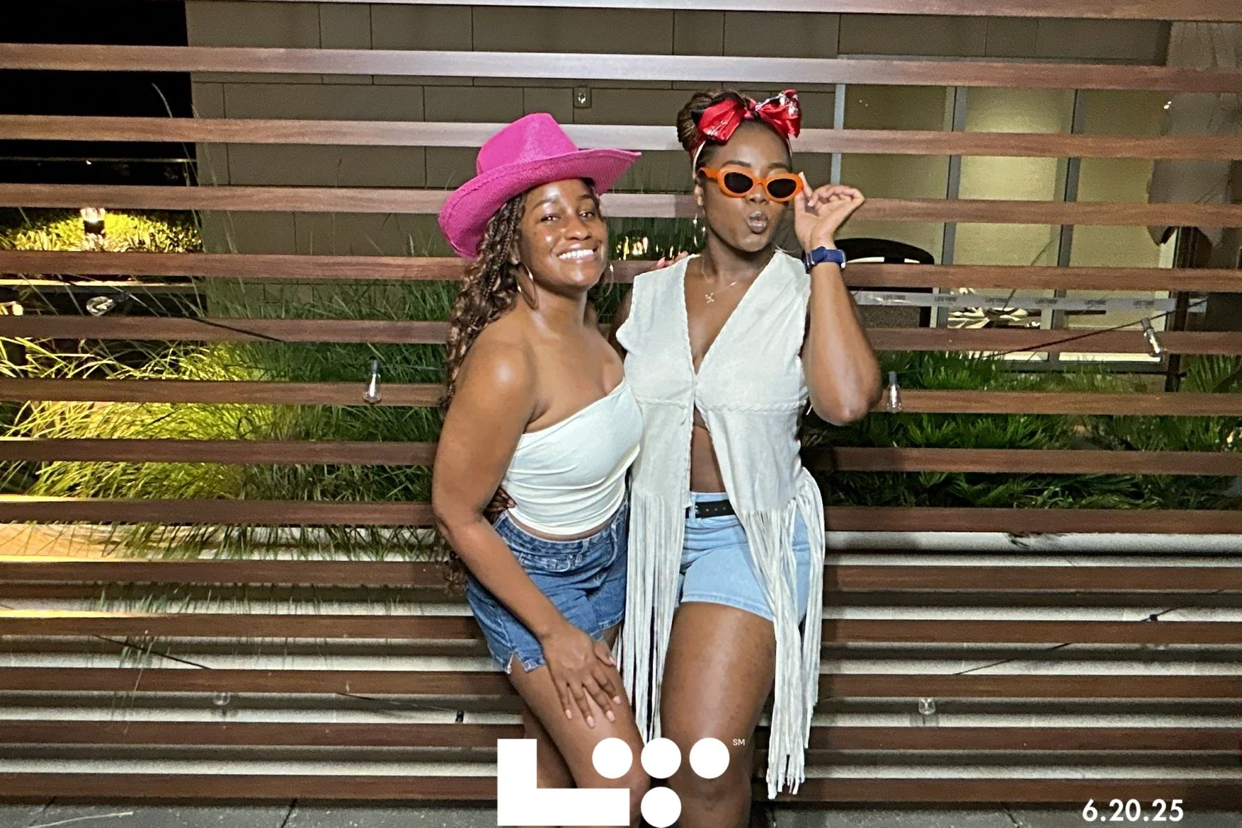 Two women posing outdoors at night in front of a wooden slatted wall with greenery, one wearing a pink hat and denim shorts, and the other wearing orange sunglasses, a headband, and a light-colored vest.
