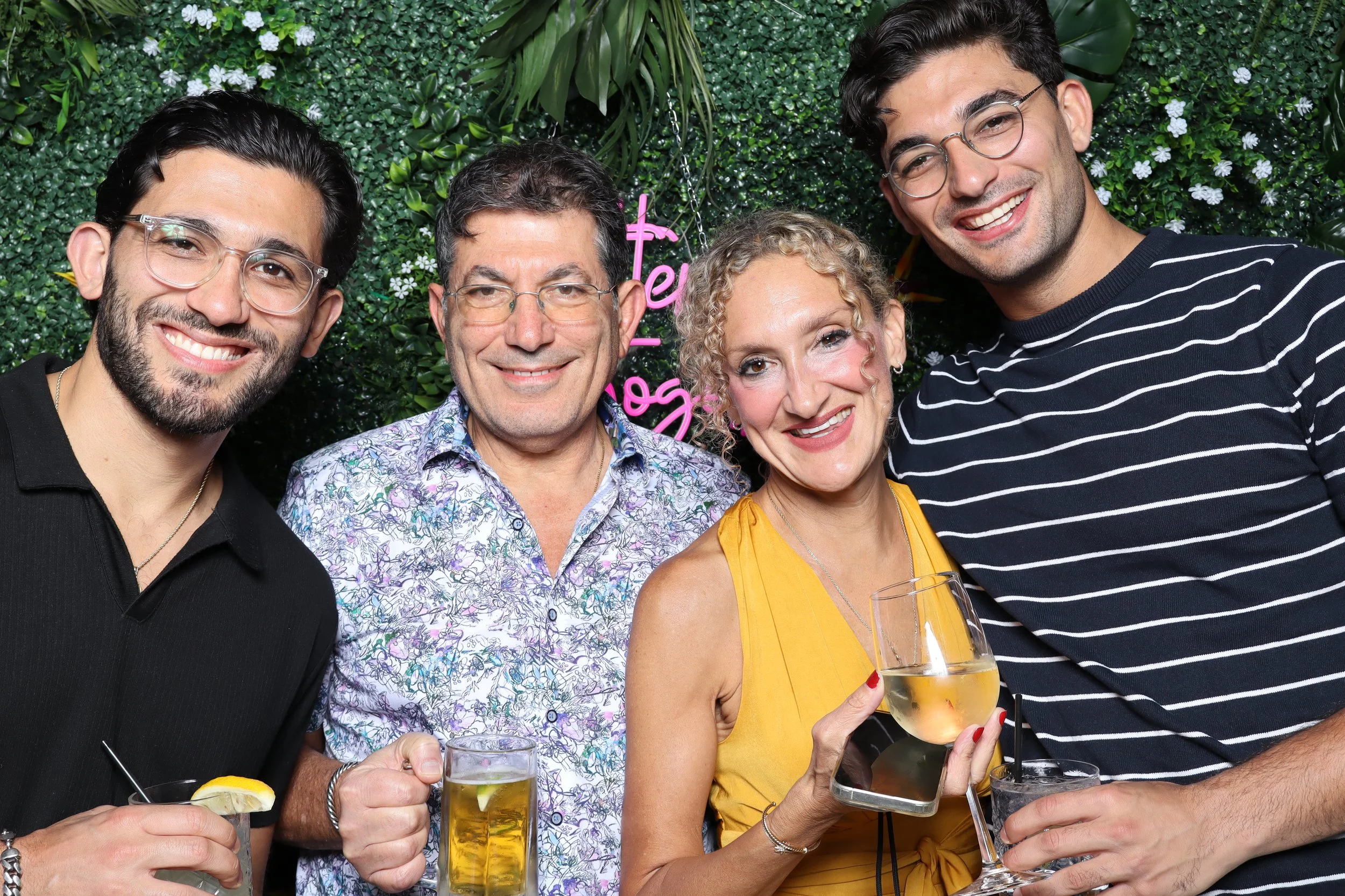 Group of five smiling people posing together at a celebration, holding drinks, with a green leafy background and a pink neon sign.