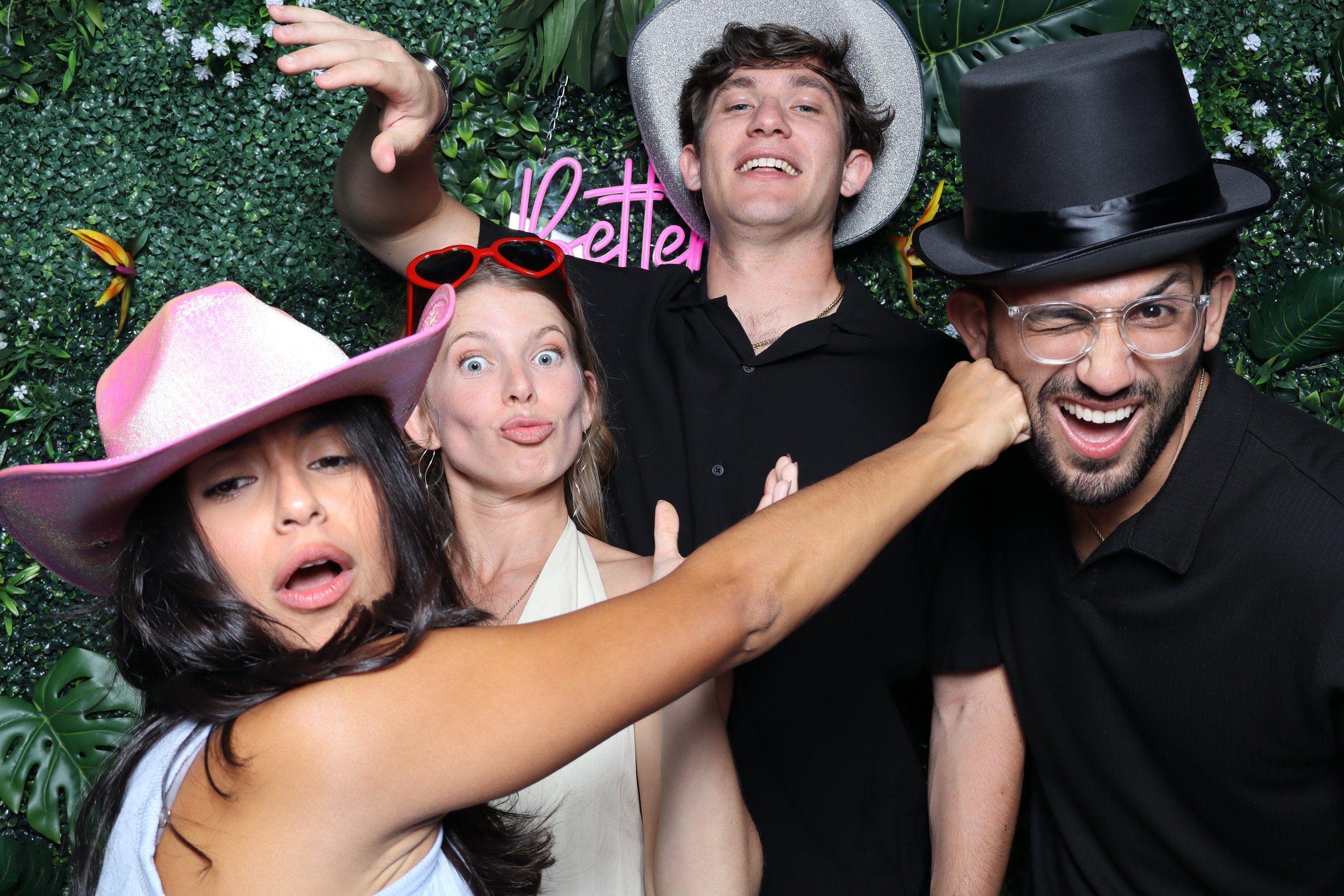 Four friends taking a playful selfie in front of a green leafy backdrop with pink neon sign reading 'Better'. They are wearing fun accessories, including a pink cowboy hat, red heart-shaped sunglasses, a silver cowboy hat, and a black top hat, and ma