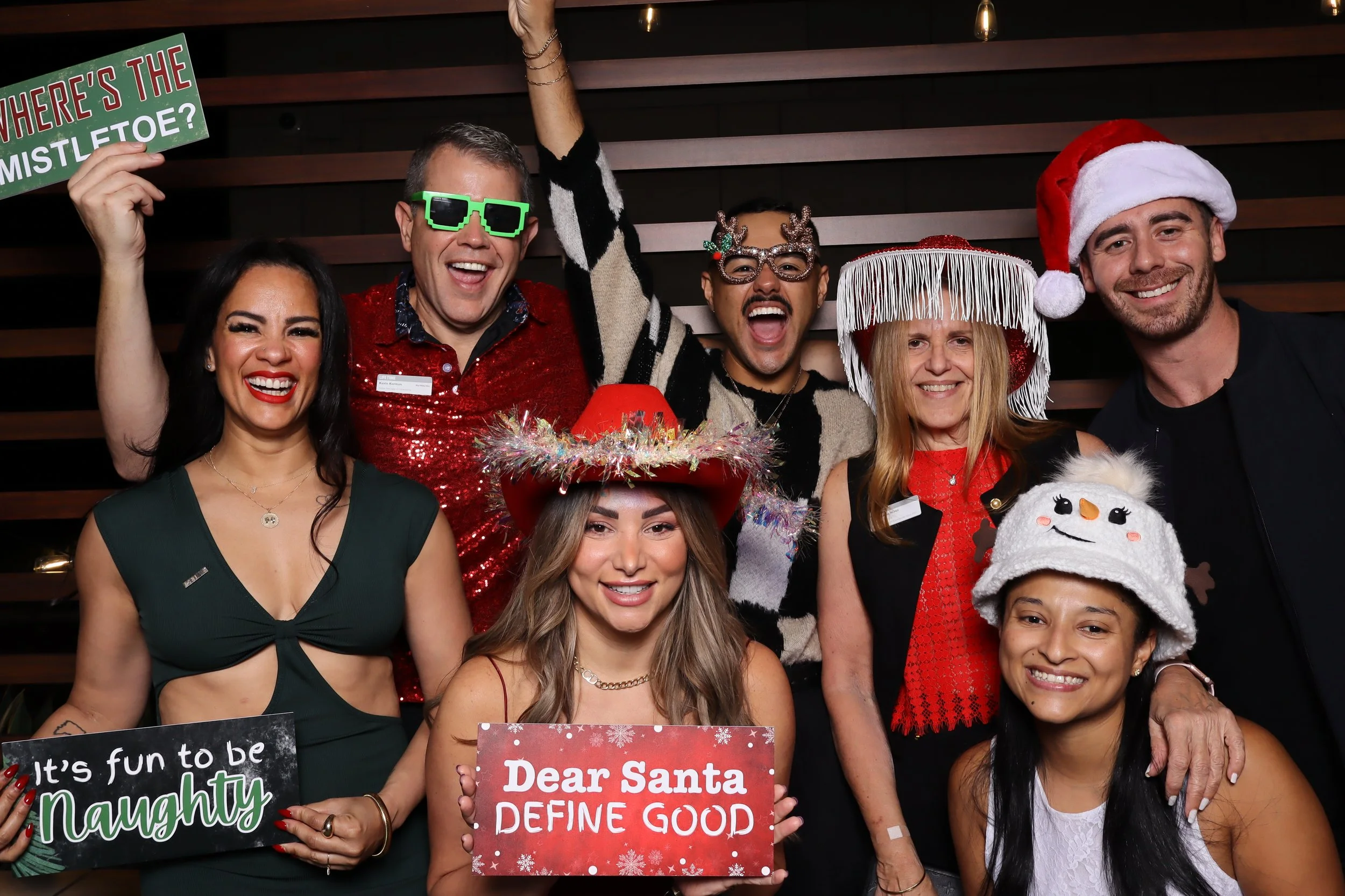 Group of people celebrating Christmas with festive hats and accessories, holding signs with holiday messages, smiling, and wearing holiday-themed costumes.