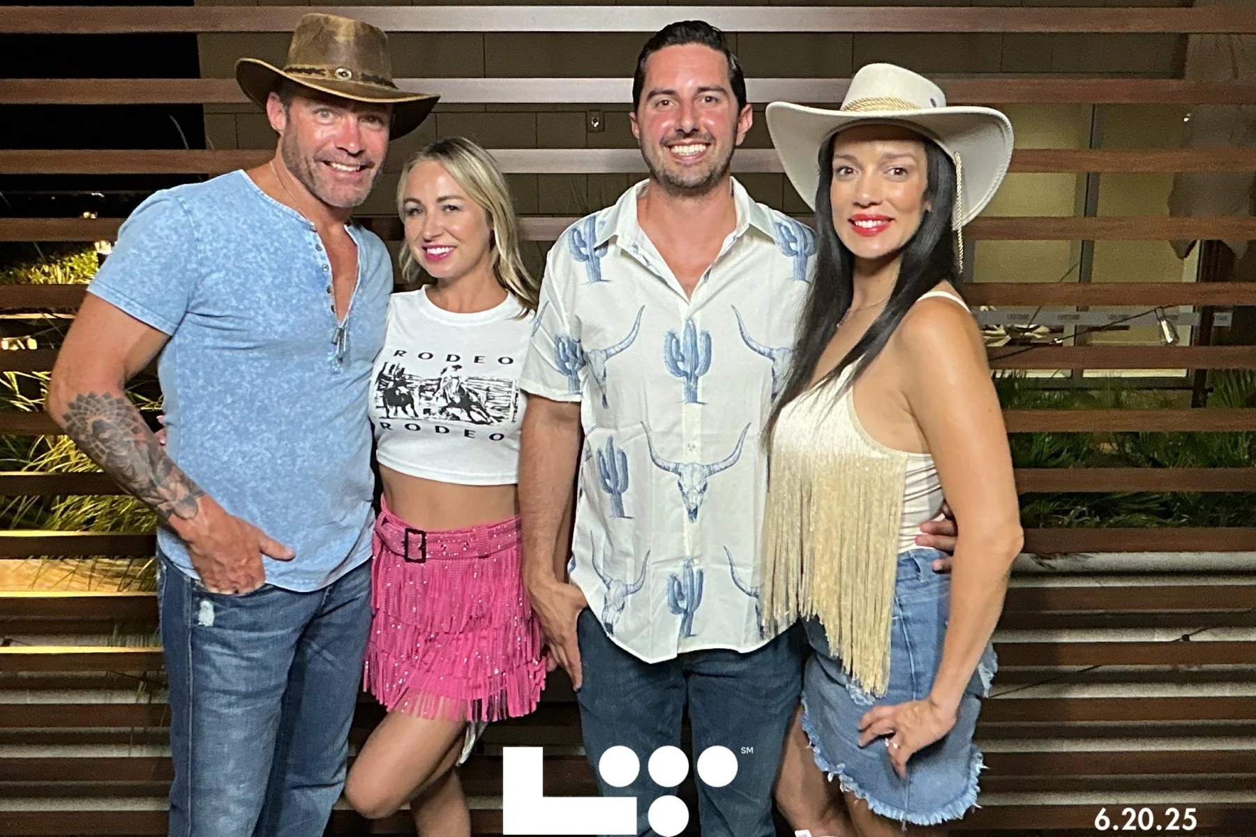 Four people smiling and posing outdoors at night, standing in front of a wooden fence. They are dressed in casual, Western-themed attire, with two women wearing hats, and one man wearing a cowboy hat. The date “6.20.25” is visible in the bottom right