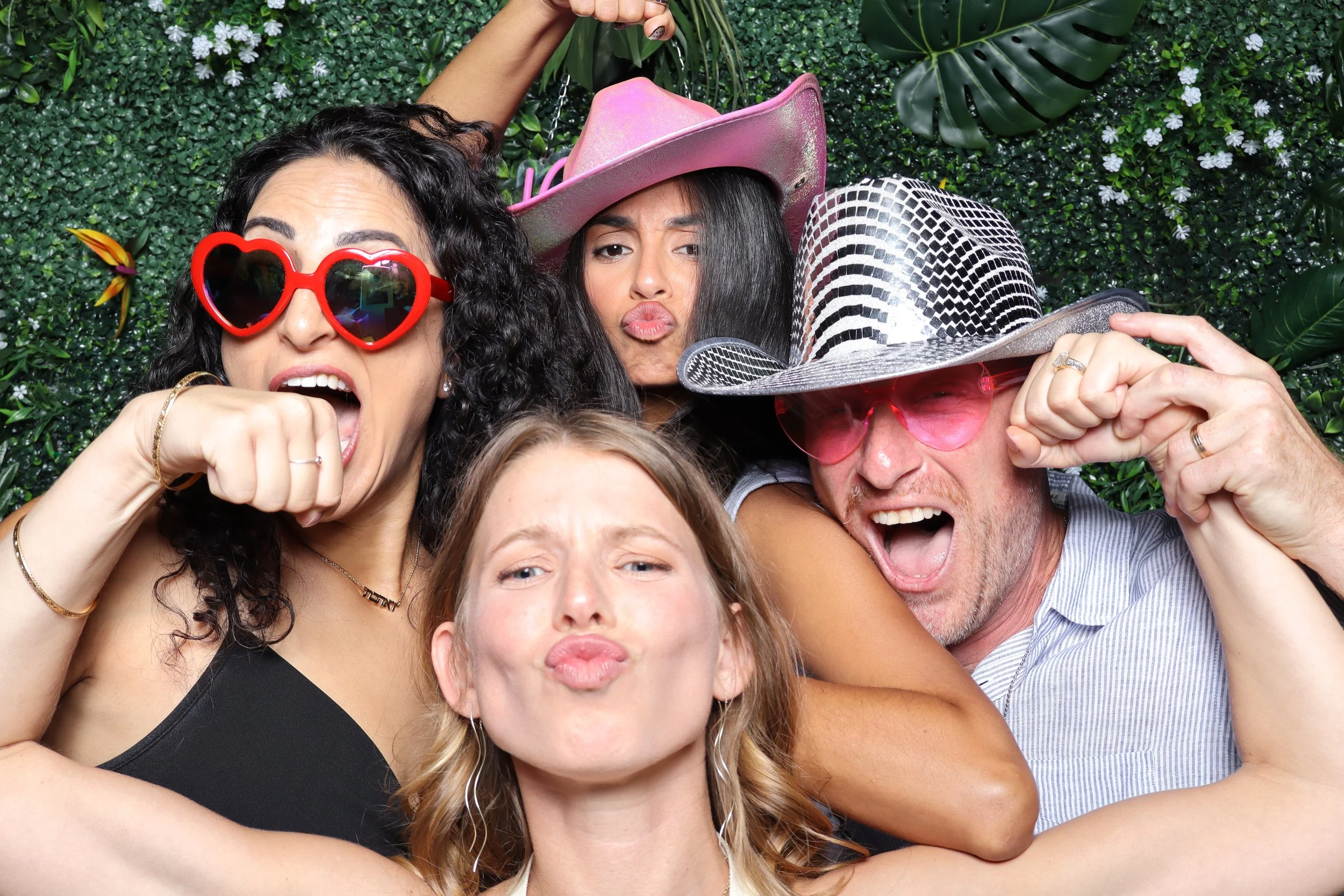 Group of four friends celebrating and posing playfully in front of a green leafy backdrop, wearing sunglasses and hats.