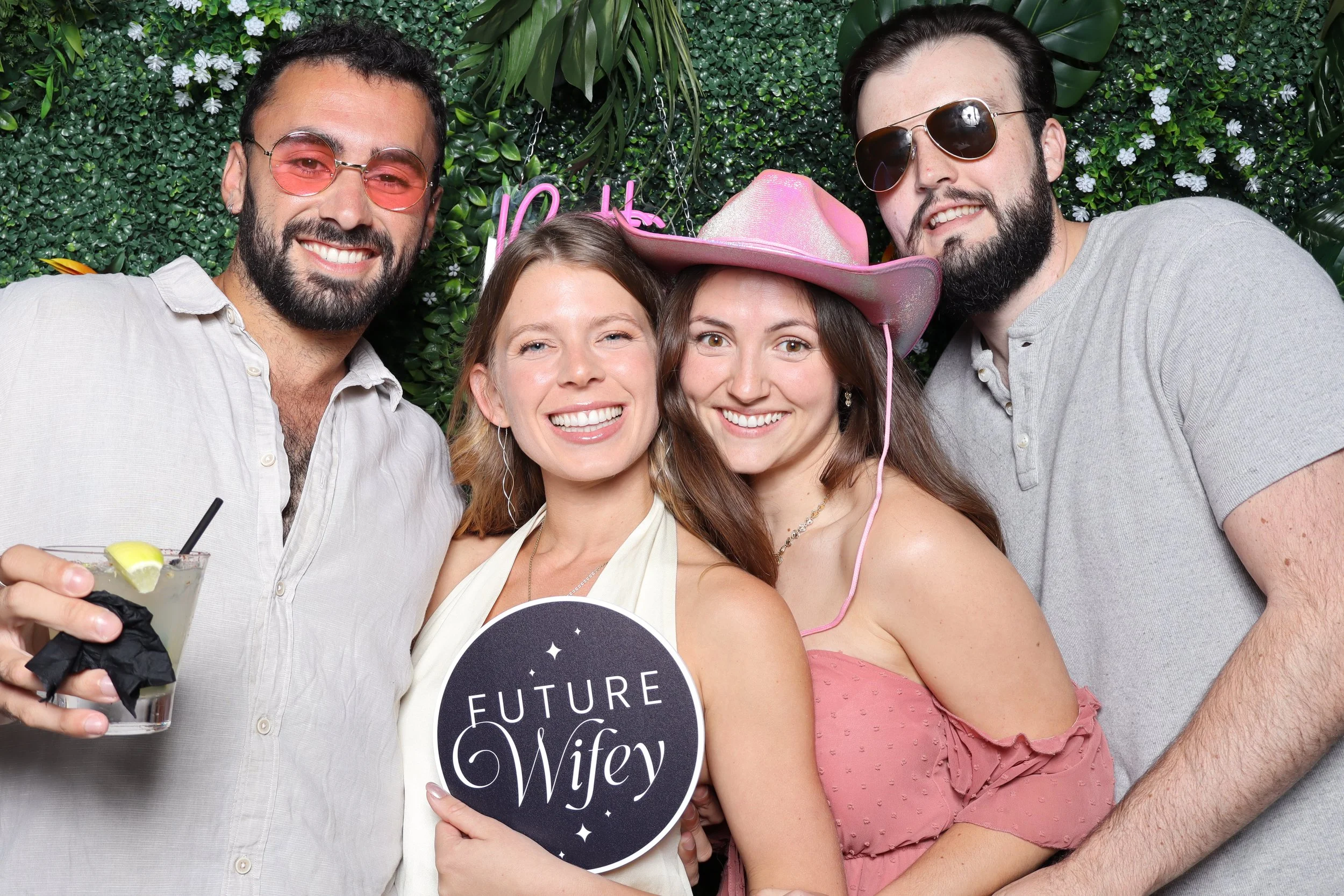 Four friends smiling at a celebration party with a green leafy backdrop. One woman holds a sign that says 'Future Wifey,' and another woman wears a pink cowboy hat. The men are wearing sunglasses, and one is holding a drink. They all appear happy and