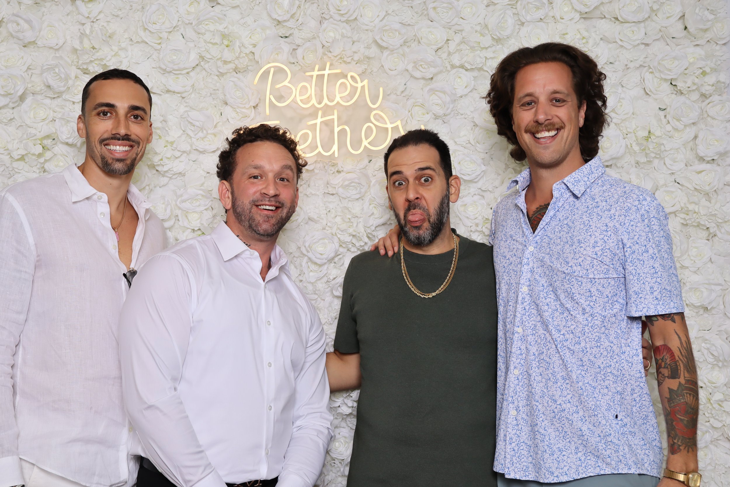 Four men standing in front of a white floral backdrop with a neon sign that reads 'Better Together.' They are smiling and posing for the photo; one of the men in the middle is making a funny face with his tongue out.