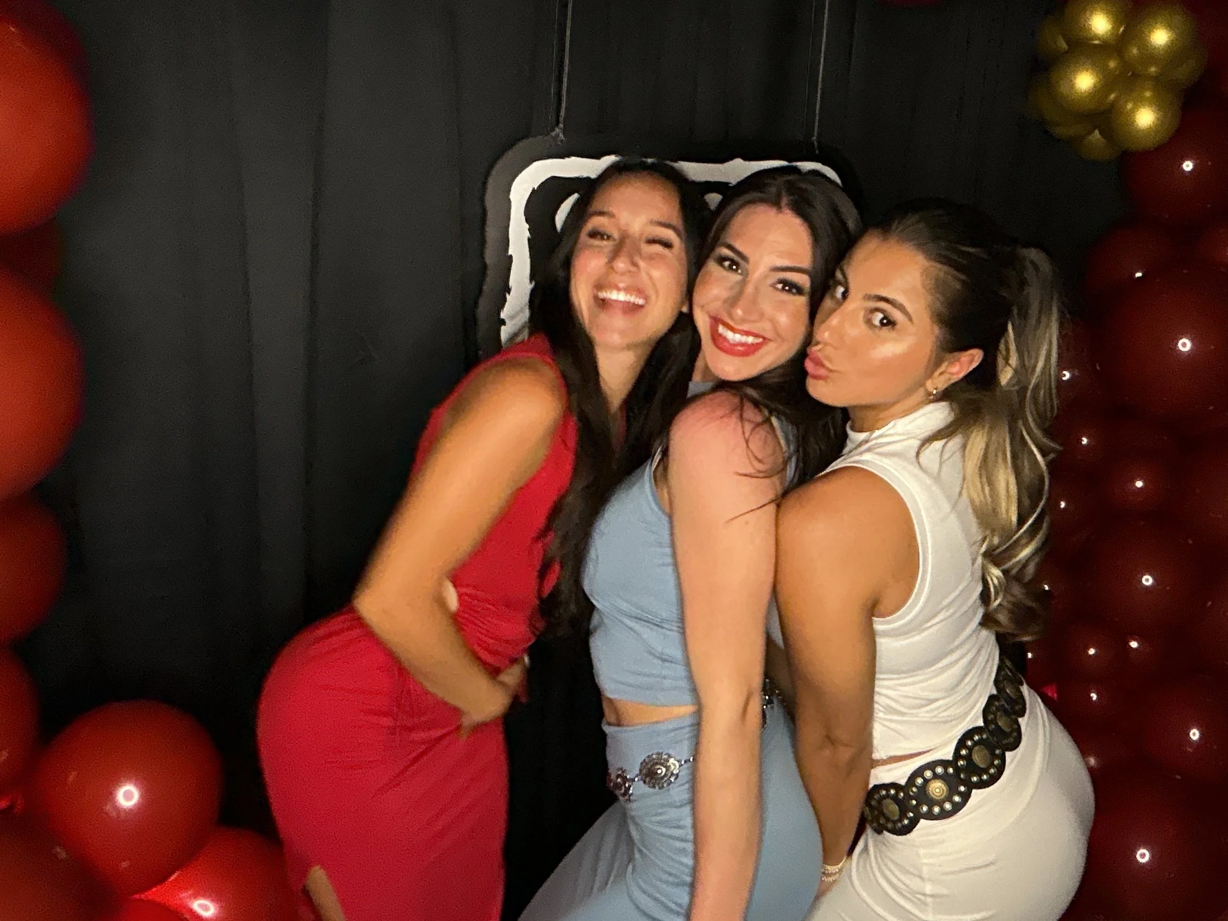 Three women at a party, smiling and posing for a photo, standing close together with balloons and a black backdrop in the background.