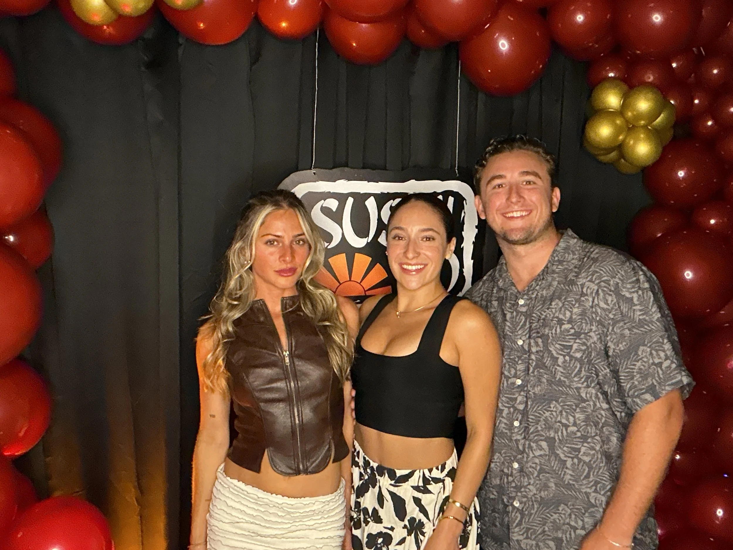 Three people posing in front of a black backdrop decorated with red and gold balloons and a sign that reads 'SUSHI'.