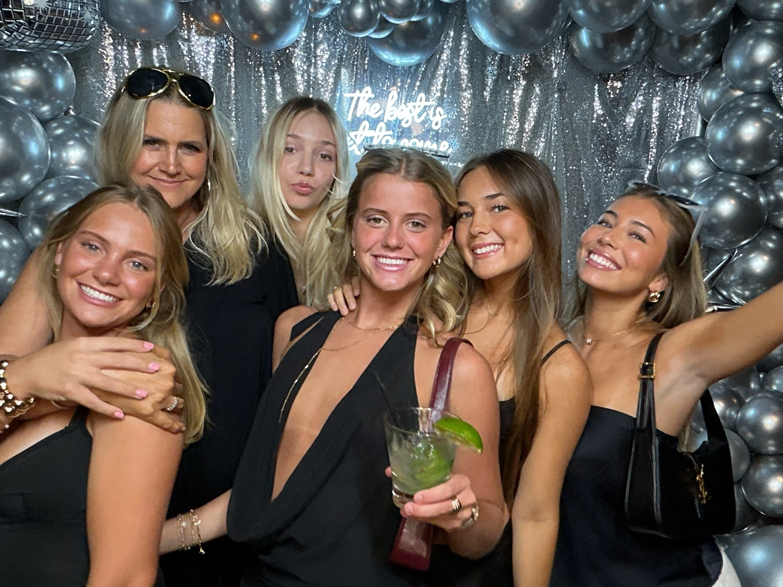 Six women at a celebration party, smiling for a selfie, with a silver balloon backdrop and a neon sign that reads 'The Boo's & The Glo'.