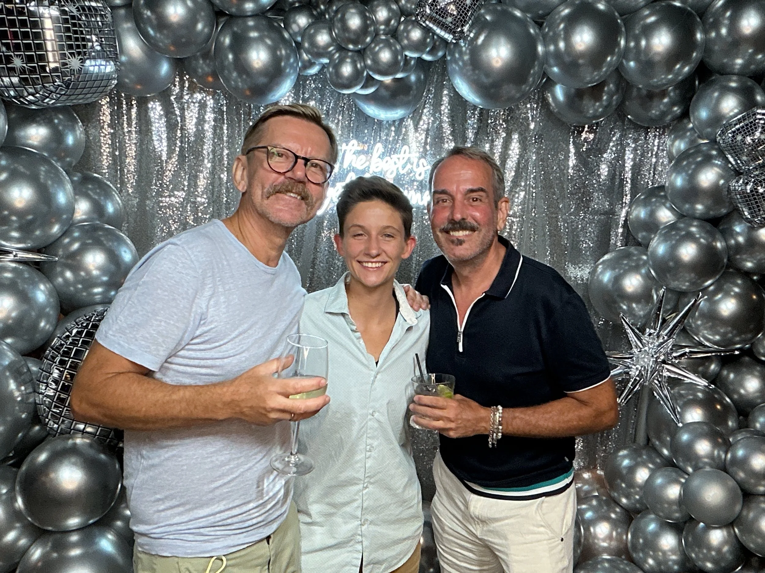 Three smiling people, two men and one woman, enjoying drinks at a celebration with silver balloons and a sparkly backdrop.