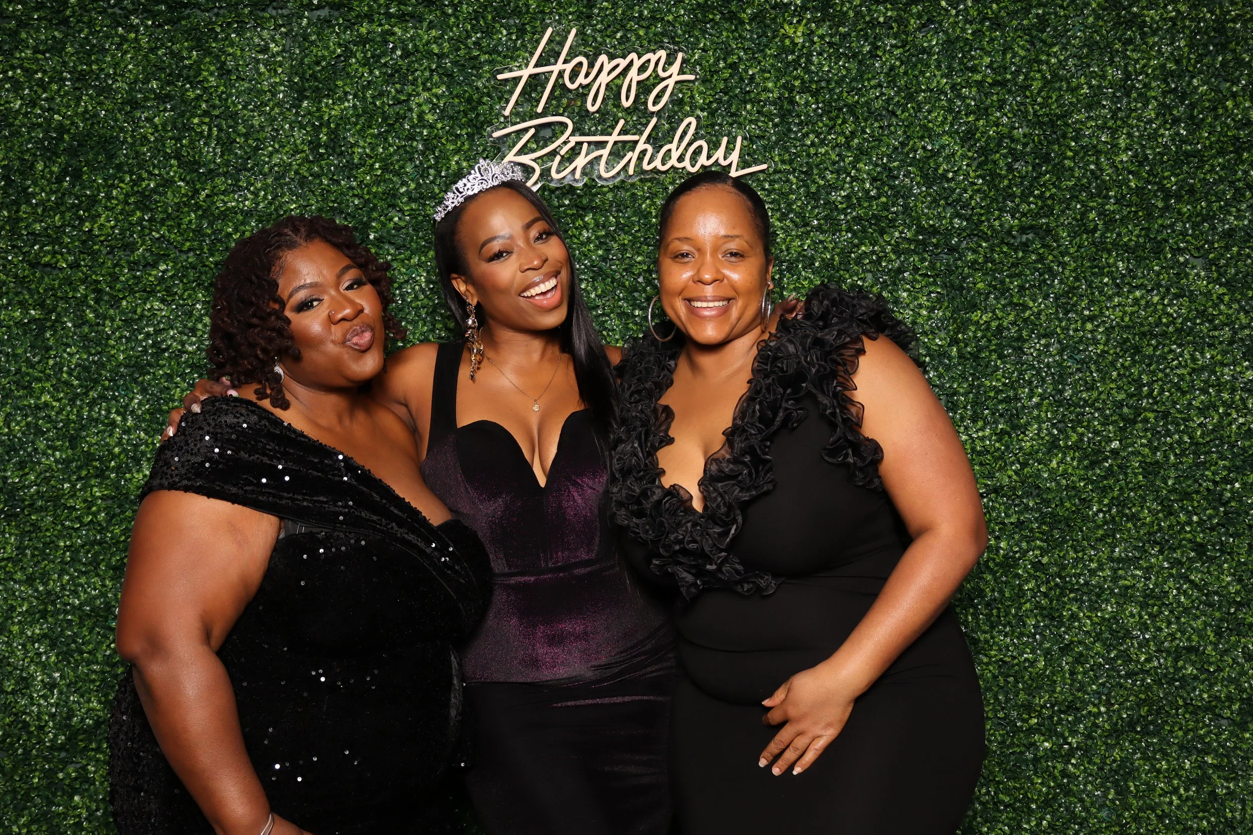 Three women dressed in black, standing close together in front of a green leafy backdrop with a neon sign that says "Happy Birthday." They are smiling and celebrating a birthday event.