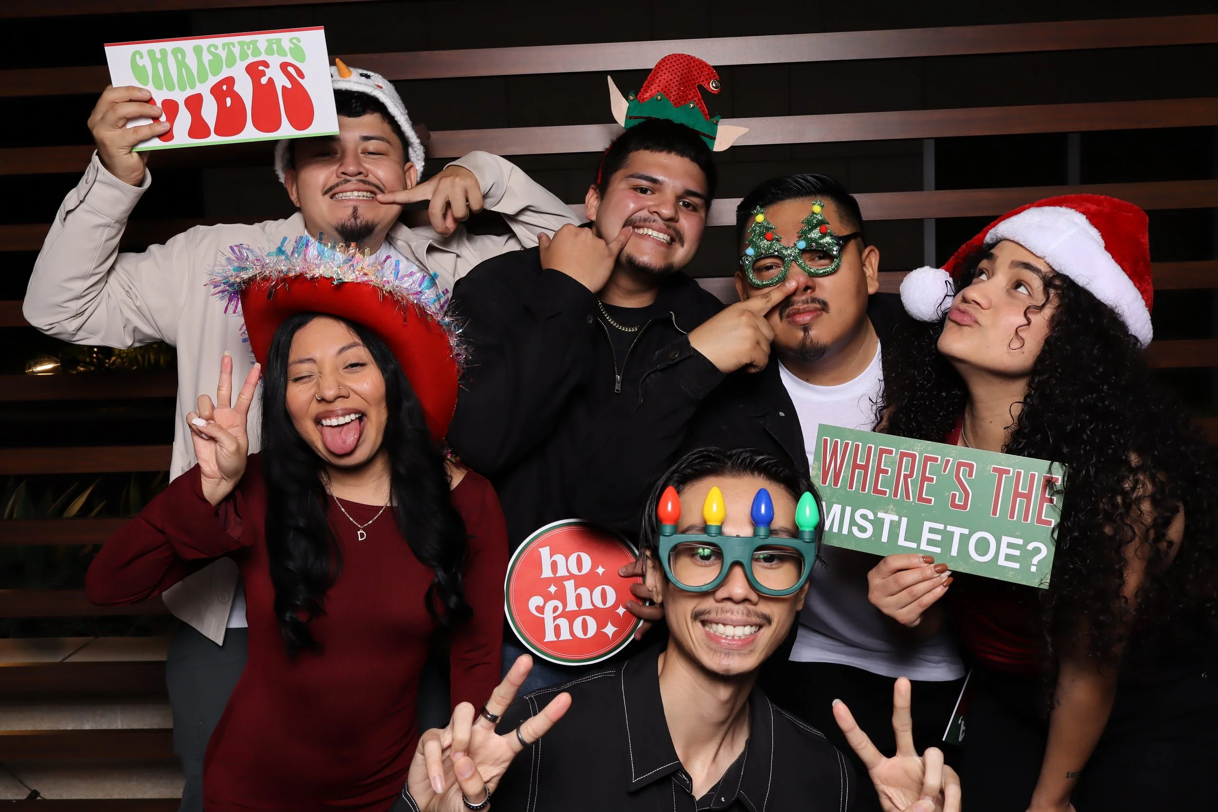 Group of six friends celebrating Christmas, wearing festive hats and accessories, holding signs that say 'Christmas Pub,' 'Ho Ho Ho,' and 'Where's the Mistletoe?' and smiling, making peace signs.