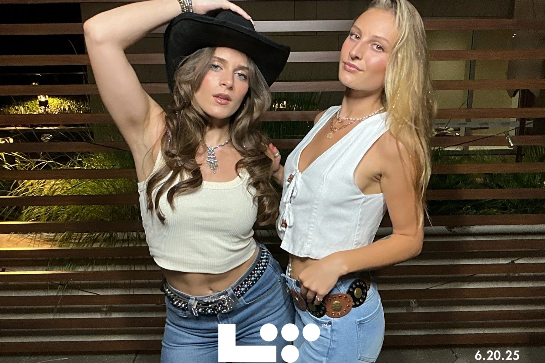 Two young women posing outdoors at night. One is wearing a black cowboy hat, a white sleeveless top, jeans, and a large belt, with long wavy brown hair. The other has light blonde hair, a white sleeveless cropped top, light blue jeans, and a decorati