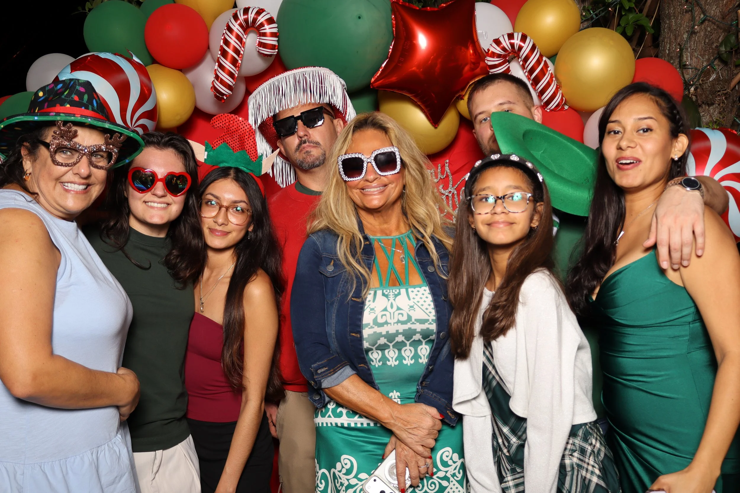 Group of people celebrating with colorful balloons and festive accessories, some wearing funny glasses, hats, and costumes, in front of a backdrop decorated for Christmas.