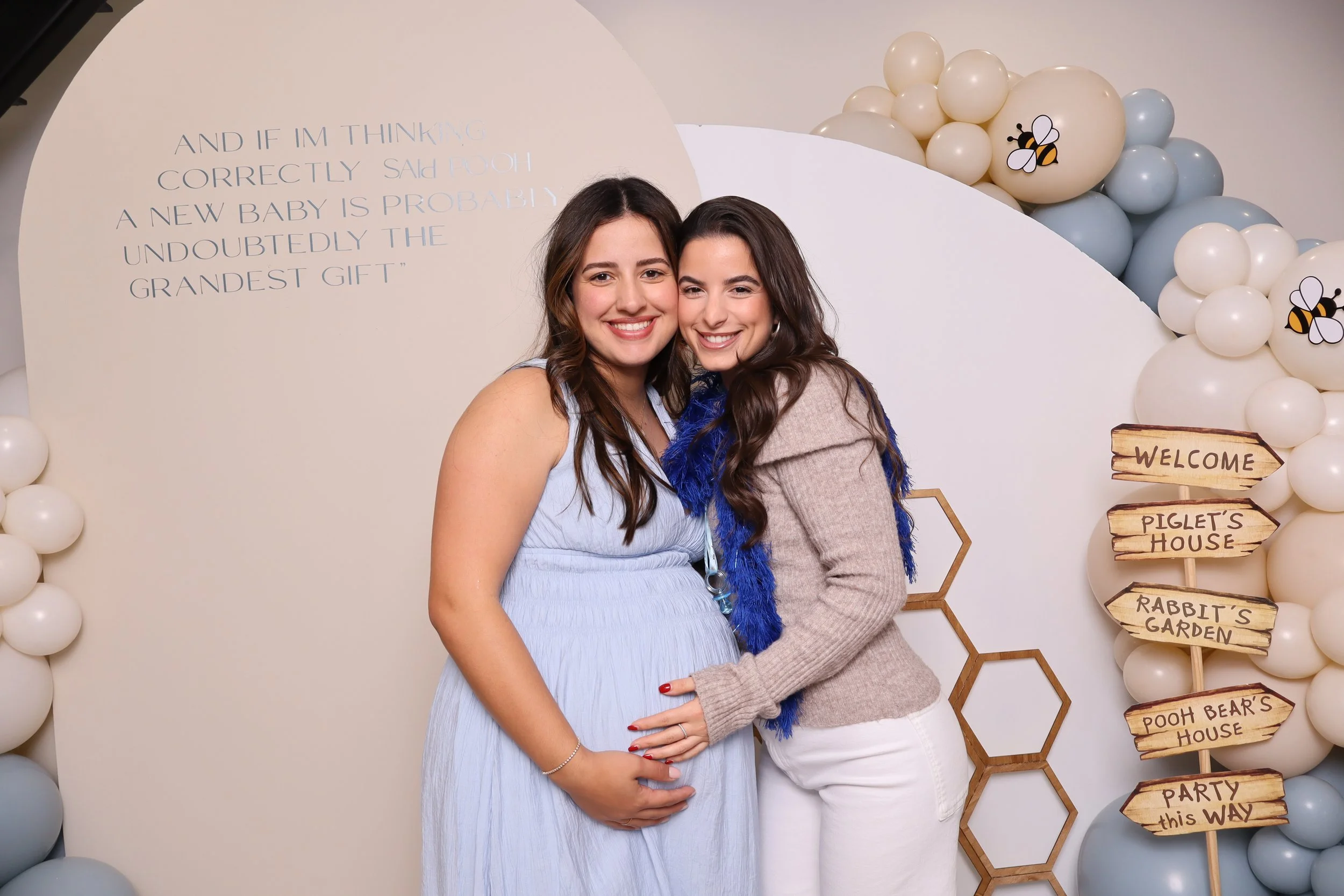 Two women smiling, with hands on pregnant woman's belly, standing in front of a decorative backdrop with balloons and a signpost with signs pointing to various themed locations.