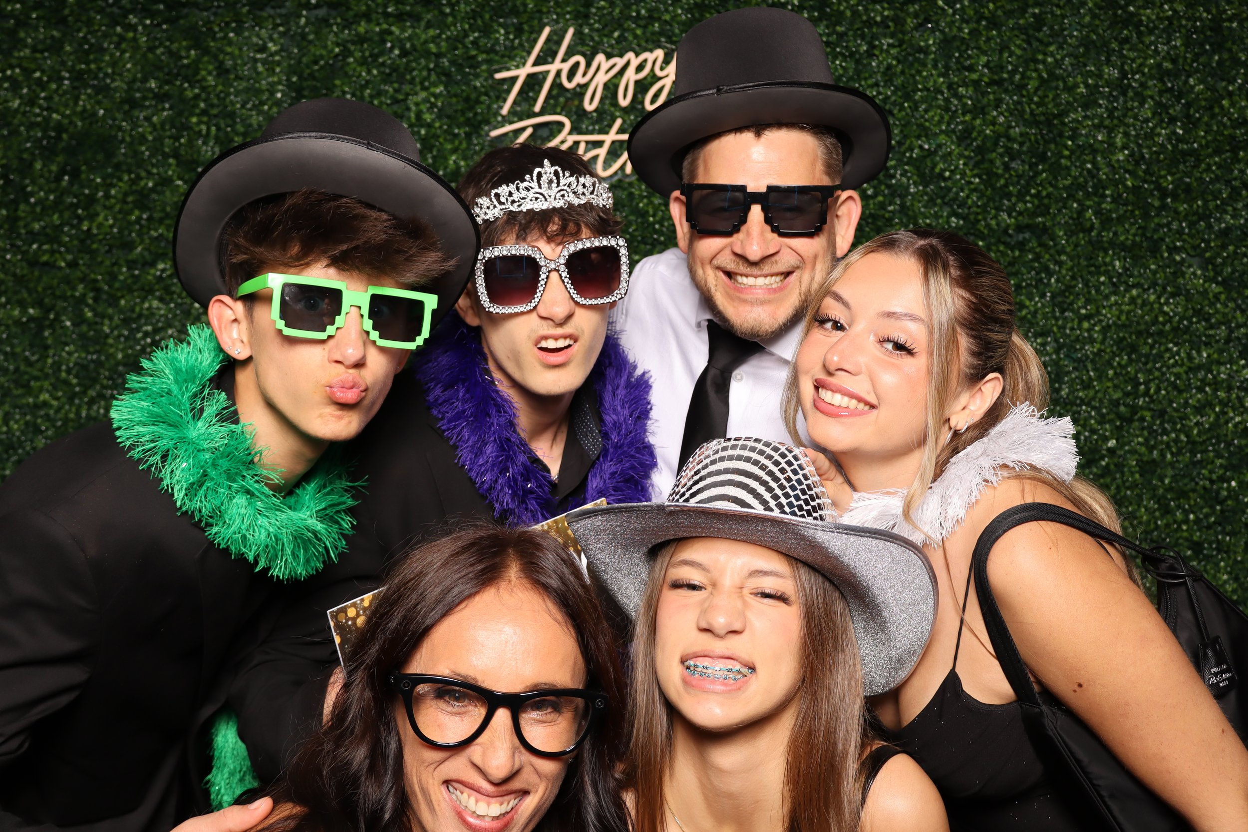 Group of friends at a birthday party posing with fun glasses, hats, and accessories in front of a green backdrop with a 'Happy Birthday' sign.