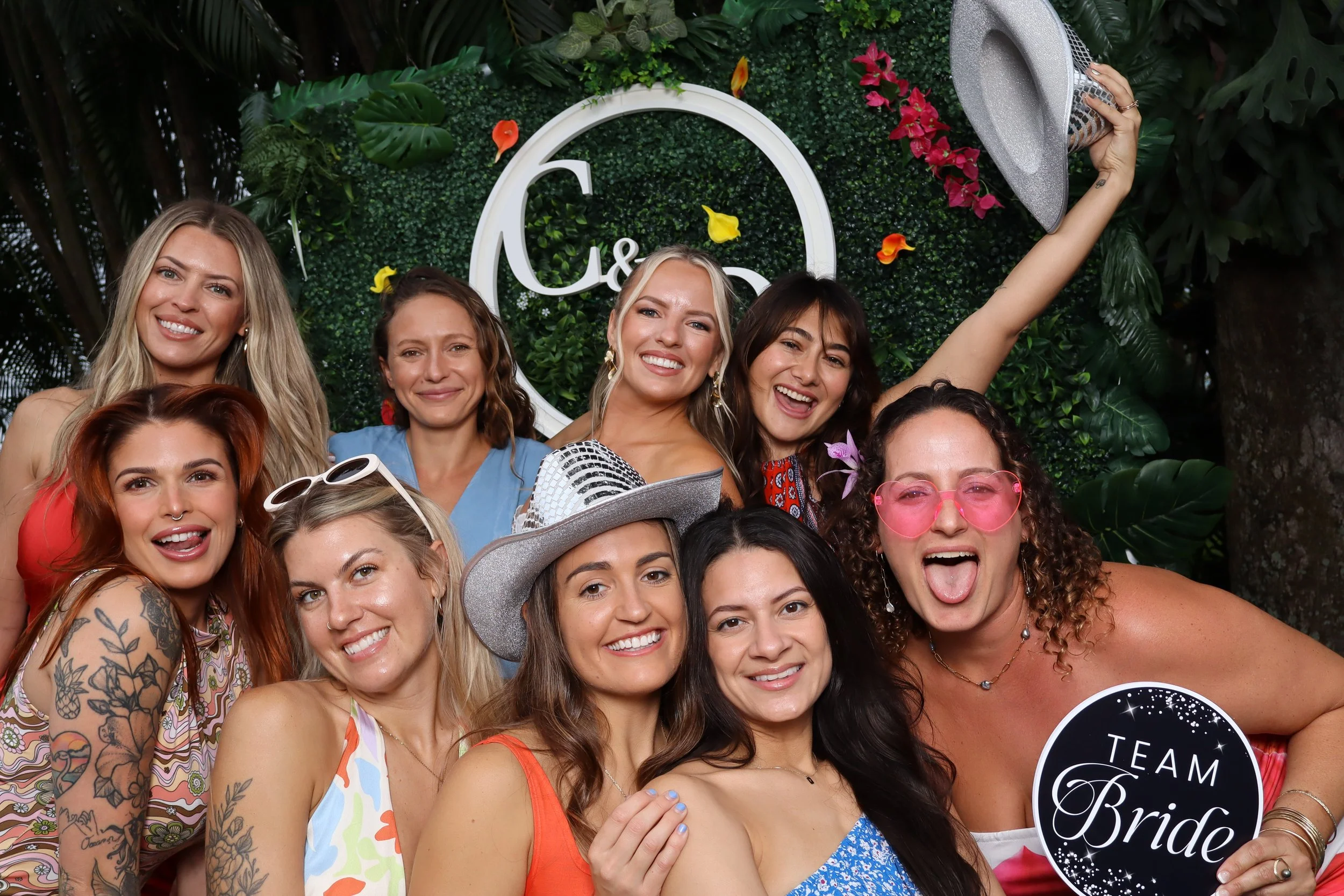A group of nine women celebrating at a bridal shower, with some wearing colorful dresses and accessories, holding a sign that reads 'TEAM Bride.' They are smiling and posing in front of a floral and greenery decorated backdrop.