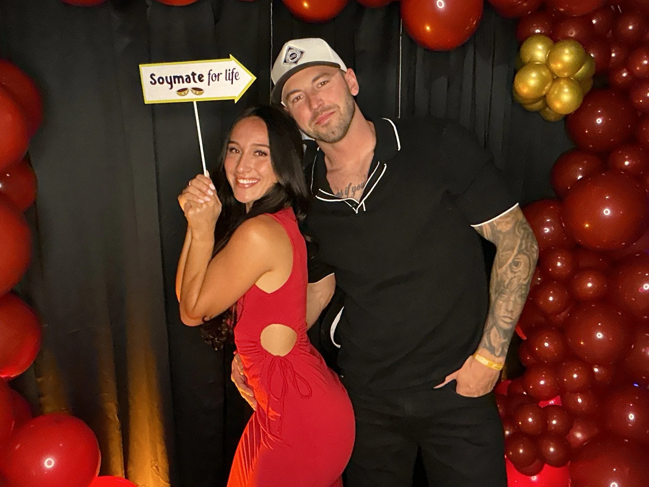 A woman in a red dress holding a sign that reads "Soymate for life," standing next to a man in a black shirt in front of a balloon backdrop of red and gold balloons.