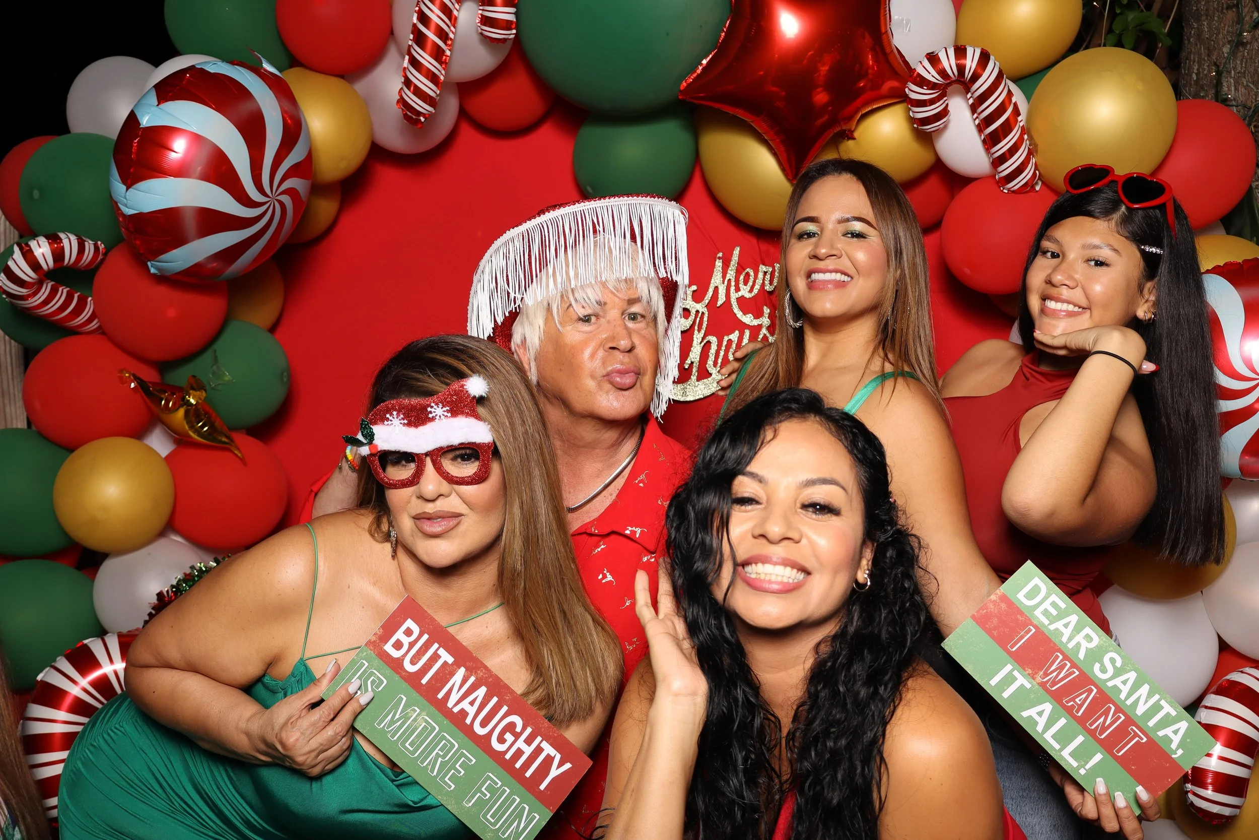 A group of five women and one man celebrating Christmas, surrounded by colorful balloons and festive decorations, some holding signs with holiday messages, and dressed in holiday-themed accessories.