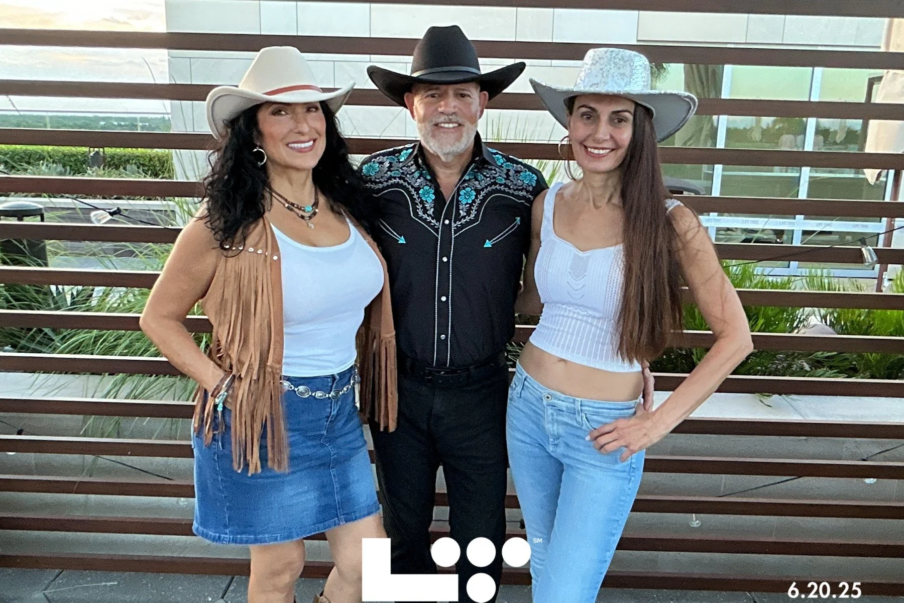 Three women and one man in cowboy hats posing together outdoors, with a modern building background. The women are wearing white tops and jeans, and the man is wearing a black Western shirt with embroidery.