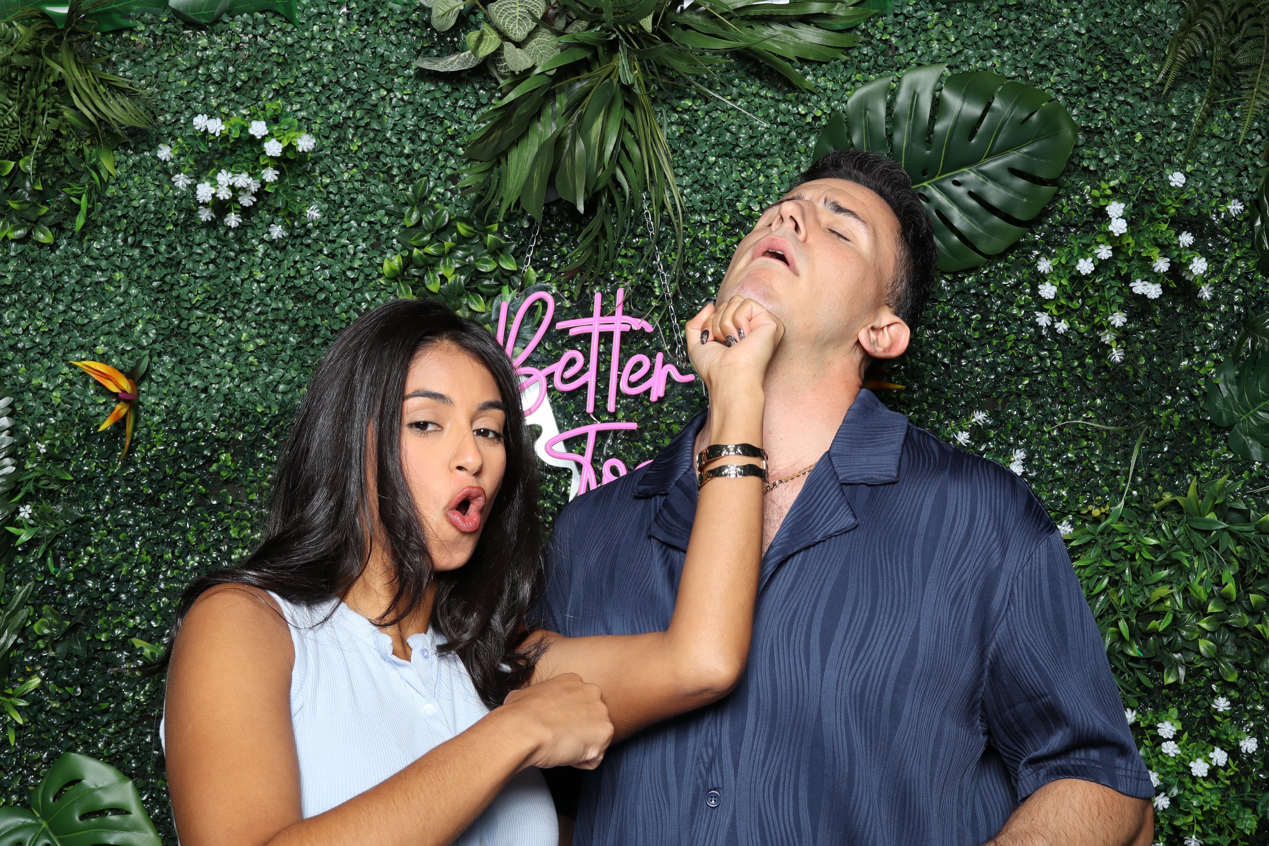 A woman playfully pinching a man's chin in front of a green leafy background with a pink neon sign that says 'Better Together'