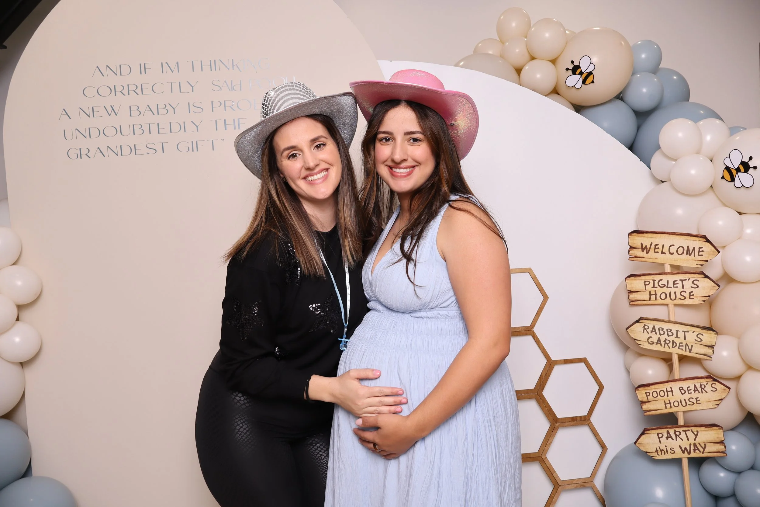 Two women, one pregnant, smiling at a party with pastel decorations and a decoration with a honeycomb pattern. One wears a sparkly silver hat, the other a pink hat. To the right, a signpost features playful signs for different house areas, such as "P