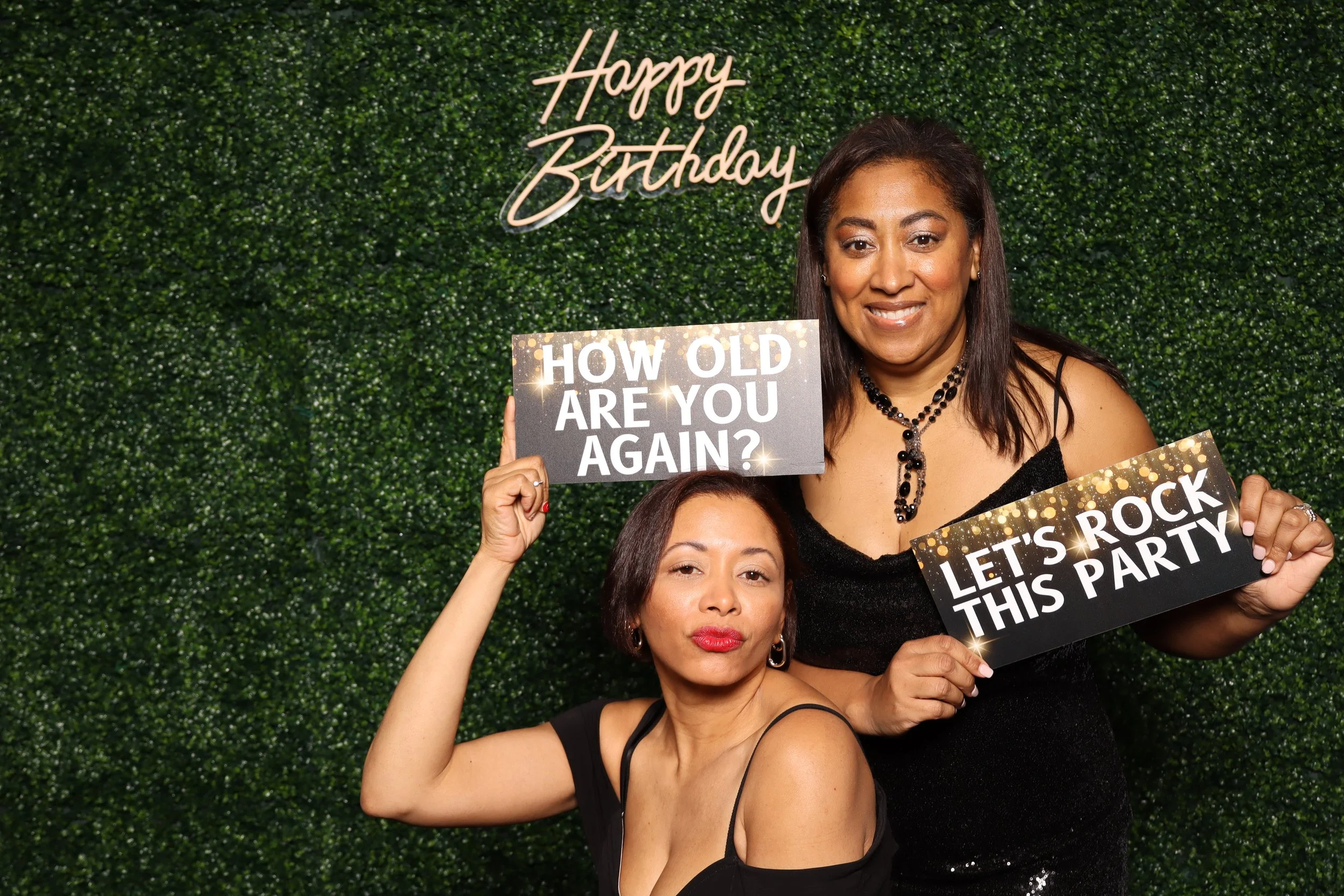 Two women posing in front of a green hedge backdrop with a 'Happy Birthday' sign, holding signs that read 'How old are you again?' and 'Let's rock this party' during a birthday celebration.