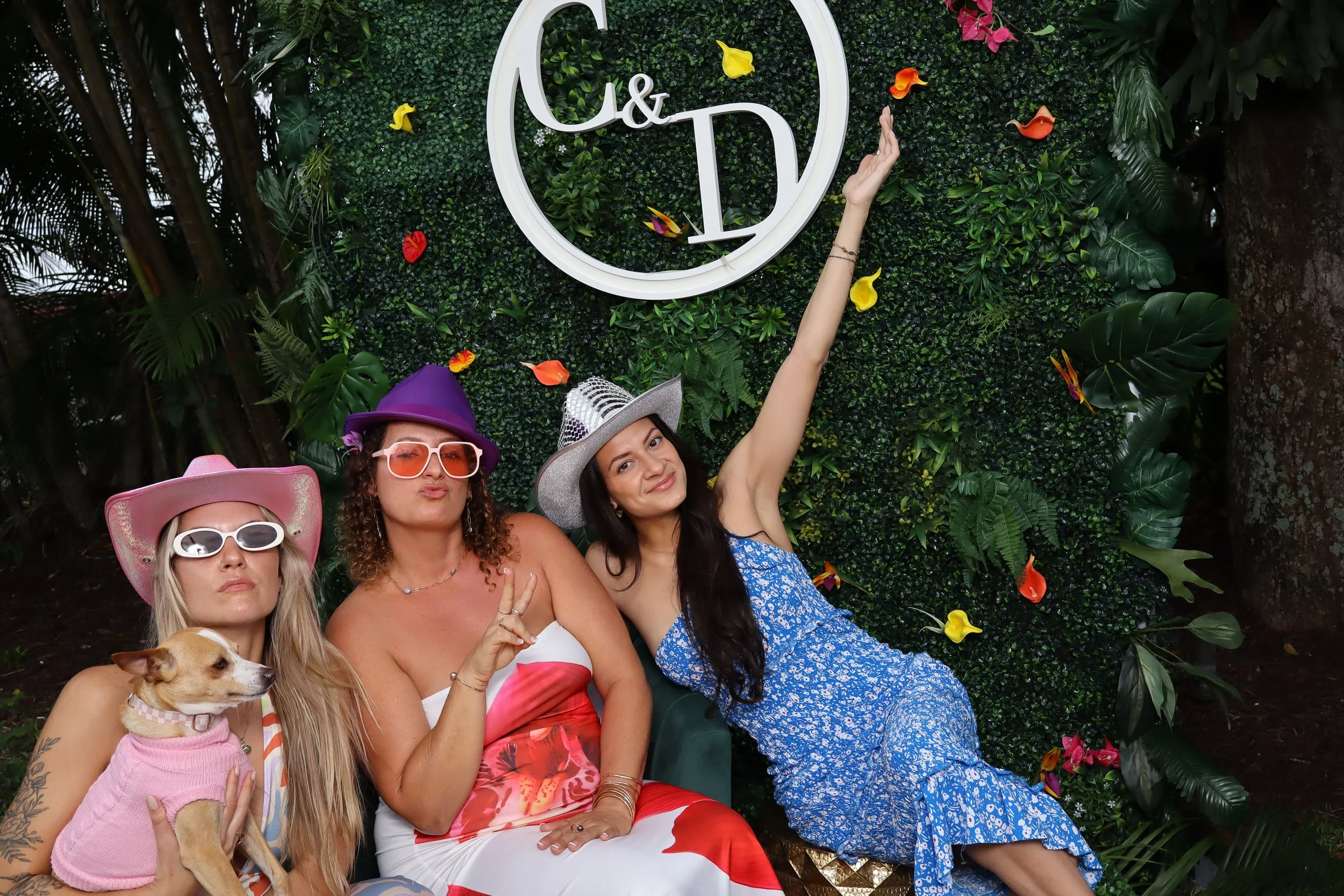 Three women and a dog posing in front of a green foliage backdrop with ornaments and a logo. The women are wearing colorful dresses, hats, and sunglasses, striking playful poses.